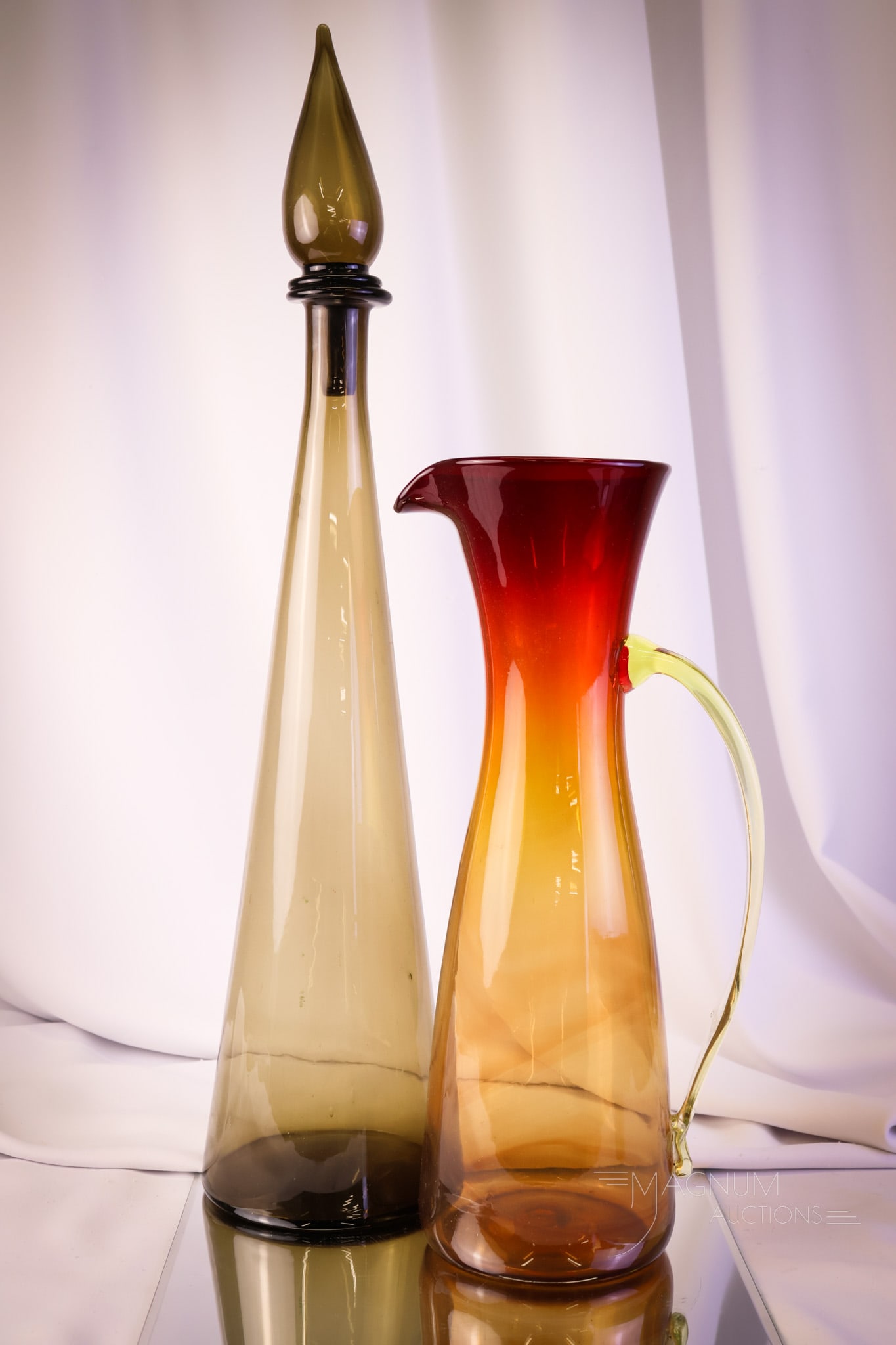 2 pc Lot MCM Empoli & Blenko Wayne Husted: A select offering of two pieces of mid-century modern art glass. This lot consists of an Empoli smoke color tall decanter along with a Blenko amberina Wayne Husted tall pitcher. The tallest measures 2