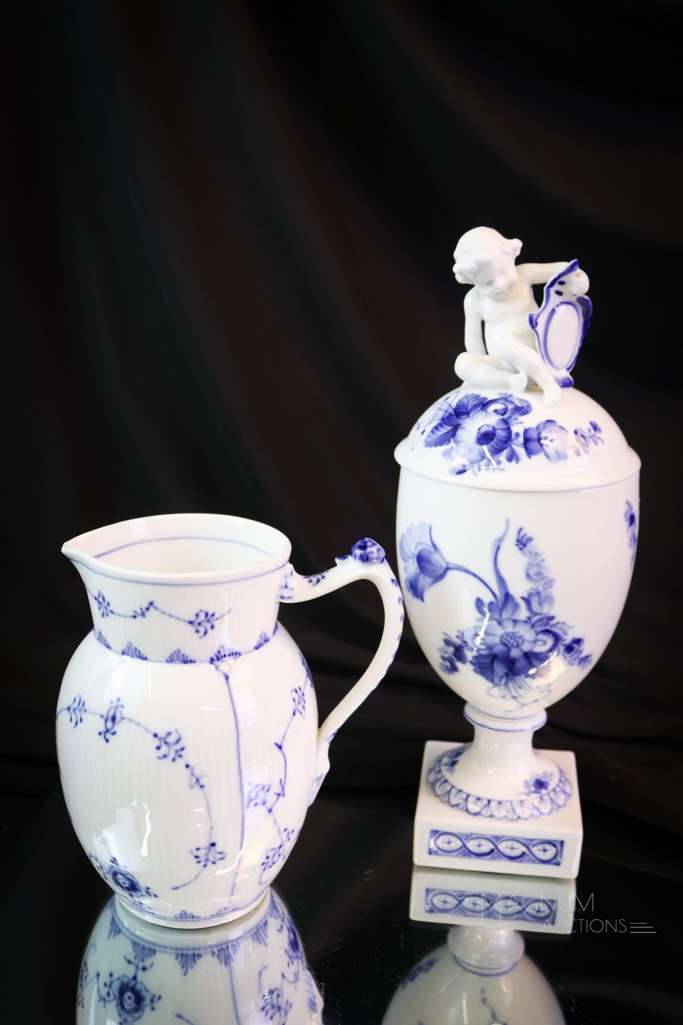 2 pc Lot Royal Copenhagen Porcelain Pitcher & Covered Urn: Two distinct finds in Royal Copenhagen porcelain. The first is a "Blue Fluted Plain" pitcher. The second piece is a "Blue Flower" covered urn with a cherub finial. Each piece of the offering is marked