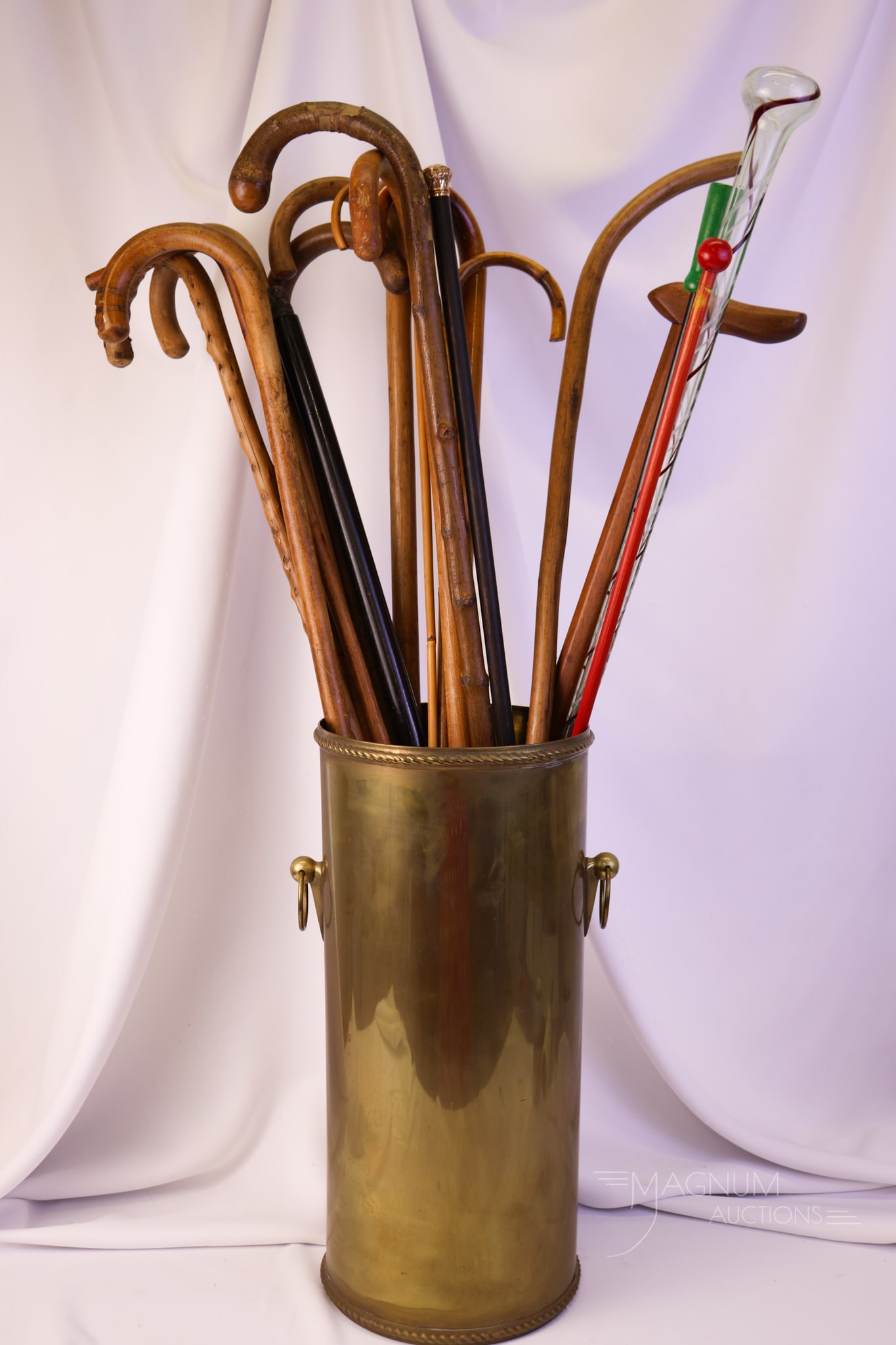 Collection of Wooden Canes Walking Sticks in Brass Umbrella Stand: A generous assortment of canes and walking sticks in a brass umbrella stand. The offering features a Victorian ebony walking stick with a gold-filled top. Two county fair sticks. There are thirteen pl