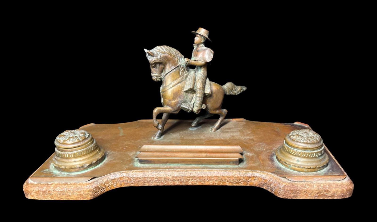 Patinated Bronze Arts and Crafts Inkwell Desk Set: This double inkwell set features a cowboy or scout on a rearing horse, flanked by two matching inkwells. There is a distinct, aged greenish-brown patina, and a groove running parallel to the horse
