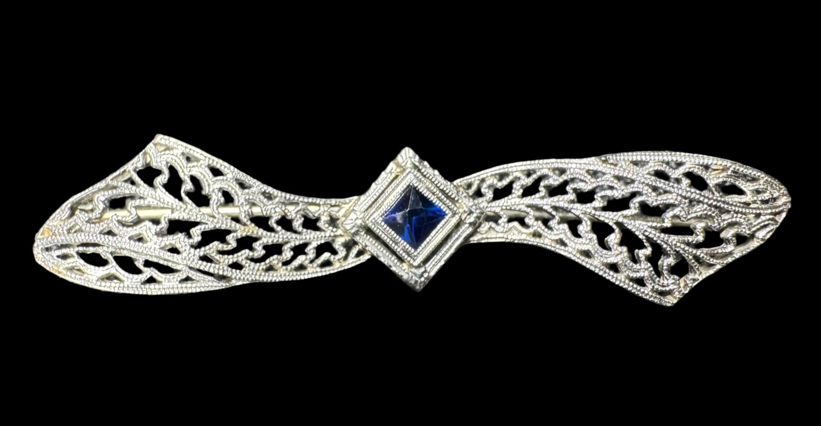 14K White Gold Art Deco Brooch: This 14K white gold Art Deco filigree brooch features a blue sapphire at the center of a “bow.” It measures approximately 2 inches wide. The weight is 2.62 grams. [PW]