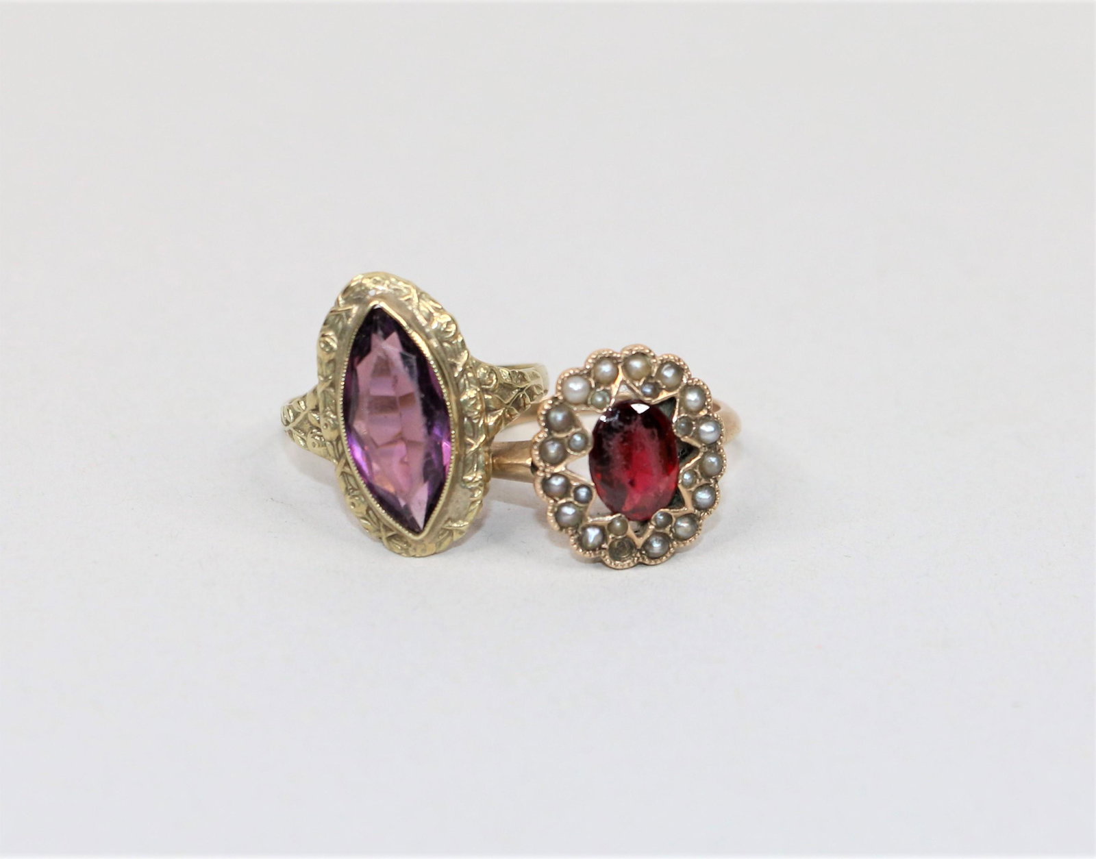 Pair of Rings: Two rings with gold tone bands; One features a purple stone and is approximately a size 6; Other ring has red colored stone surrounded by miniature pearl-like stones and is approximately a size 7; No 