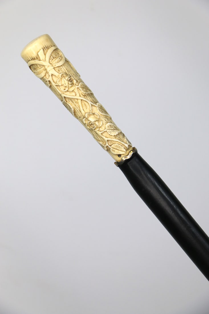 Bone Handled Cane: Antique walking stick with highly carved bone handle, sterling silver collar and ebonized wood shaft; Handle is carved with a rose motif with a crown and M on handle top; Measures approximately H 40 3