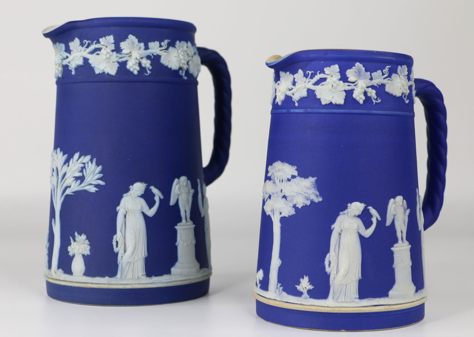 Wedgwood, Cobalt Blue Jasperware: Two Wedgwood jugs / pitchers in different sizes; Both have an impressed "Wedgwood, England" on the base; Tallest measures approximately H 6 1/2 inches; Largest size has a crack on the spout; Smaller j