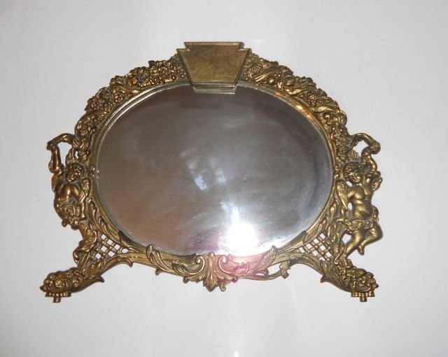 Victorian Brass Oval Table Easel Mirror.: H - 13", W - 17". Oval format mirror with elaborate relief frame of frolicking Putti's.