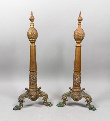 Pair of Bronze Andirons: Bronze andirons with carved designs mounted on footed base with lions head in the center. 10" L x 11" W x 25 1/8" H