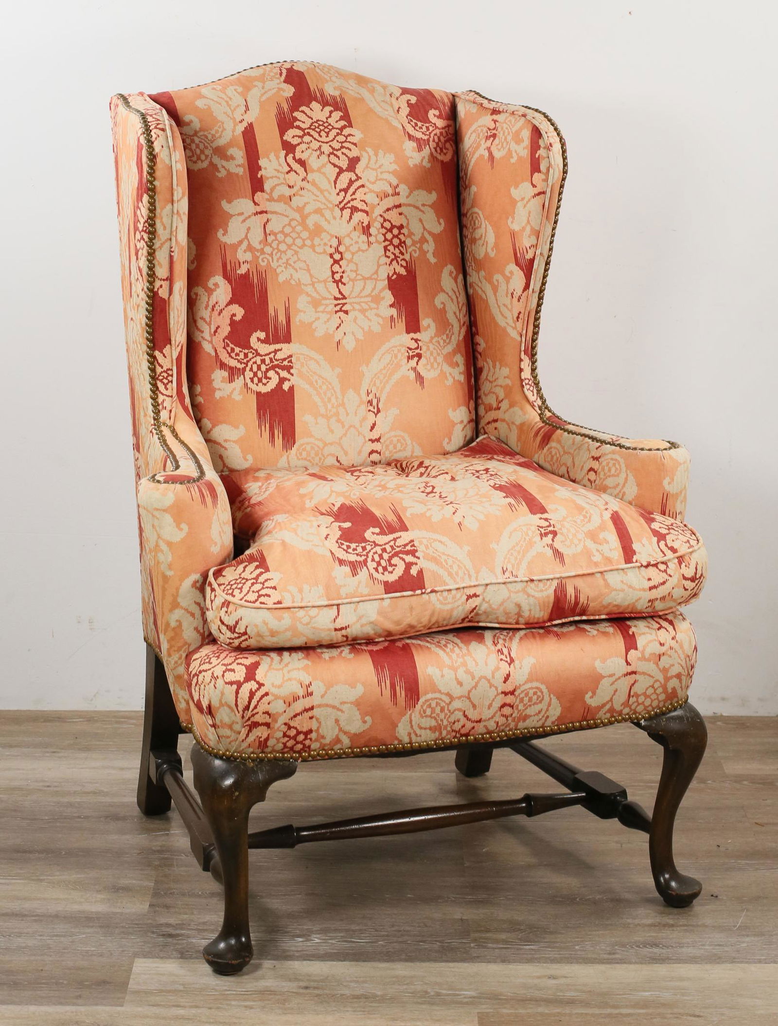 Queen Anne Style Upholstered Wingback Chair: Queen Anne Style upholstered wingback chair. American, Early 20th Century. Cabriole legs and pad feet, carved stretcher, red and salmon upholstery with brass nail head trim. 45" H x 24" D x 31 3/4" W