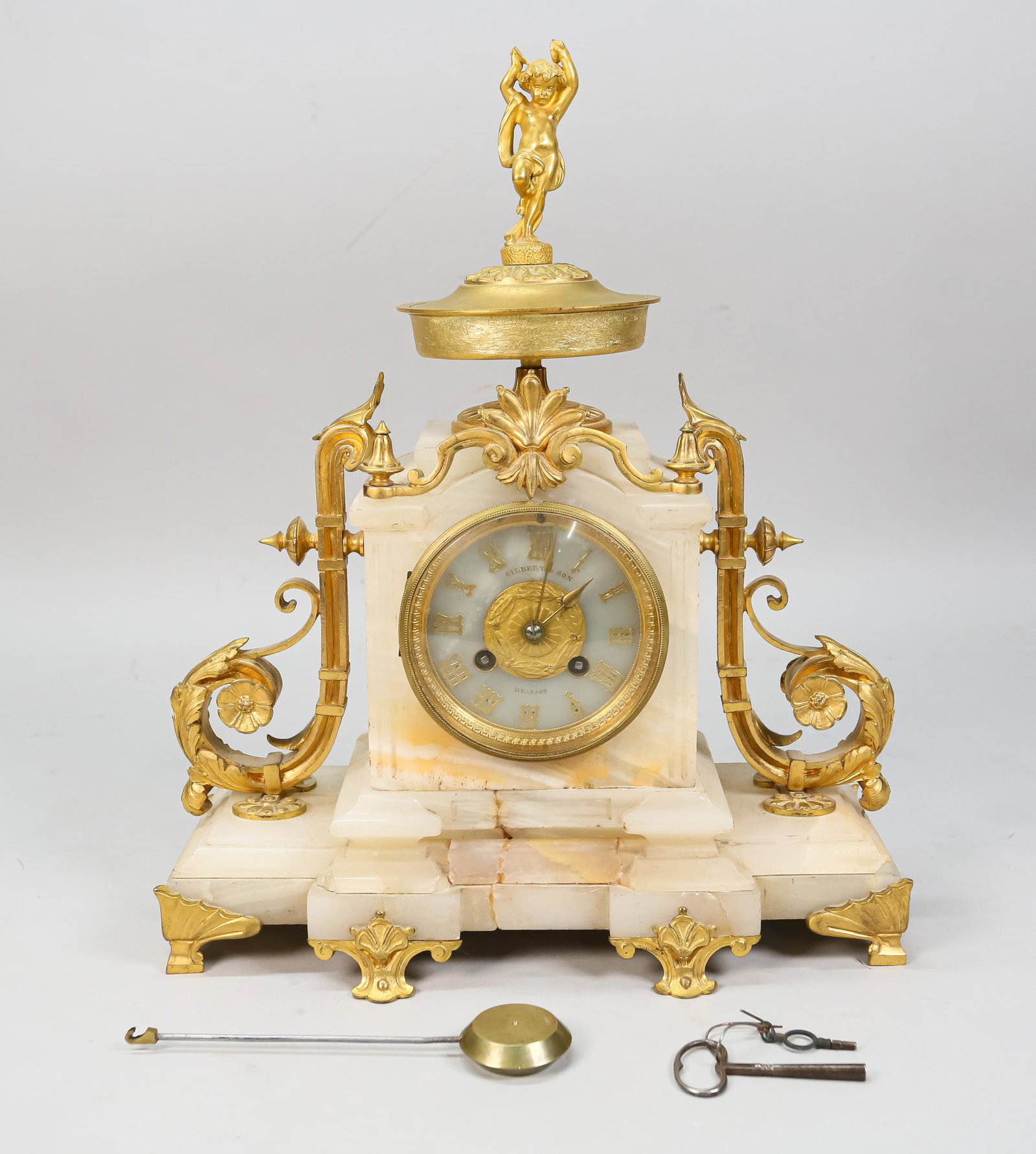Gilbert & Son Belfast Alabaster & Gilt Metal Clock: Gilbert & Son Belfast alabaster and gilt metal mantle clock. French style. Figural top with putto, mounted gilt flourishes. With key and pendulum. Loss to stone along the base. 12" L x 4 1/4" W x 14 1