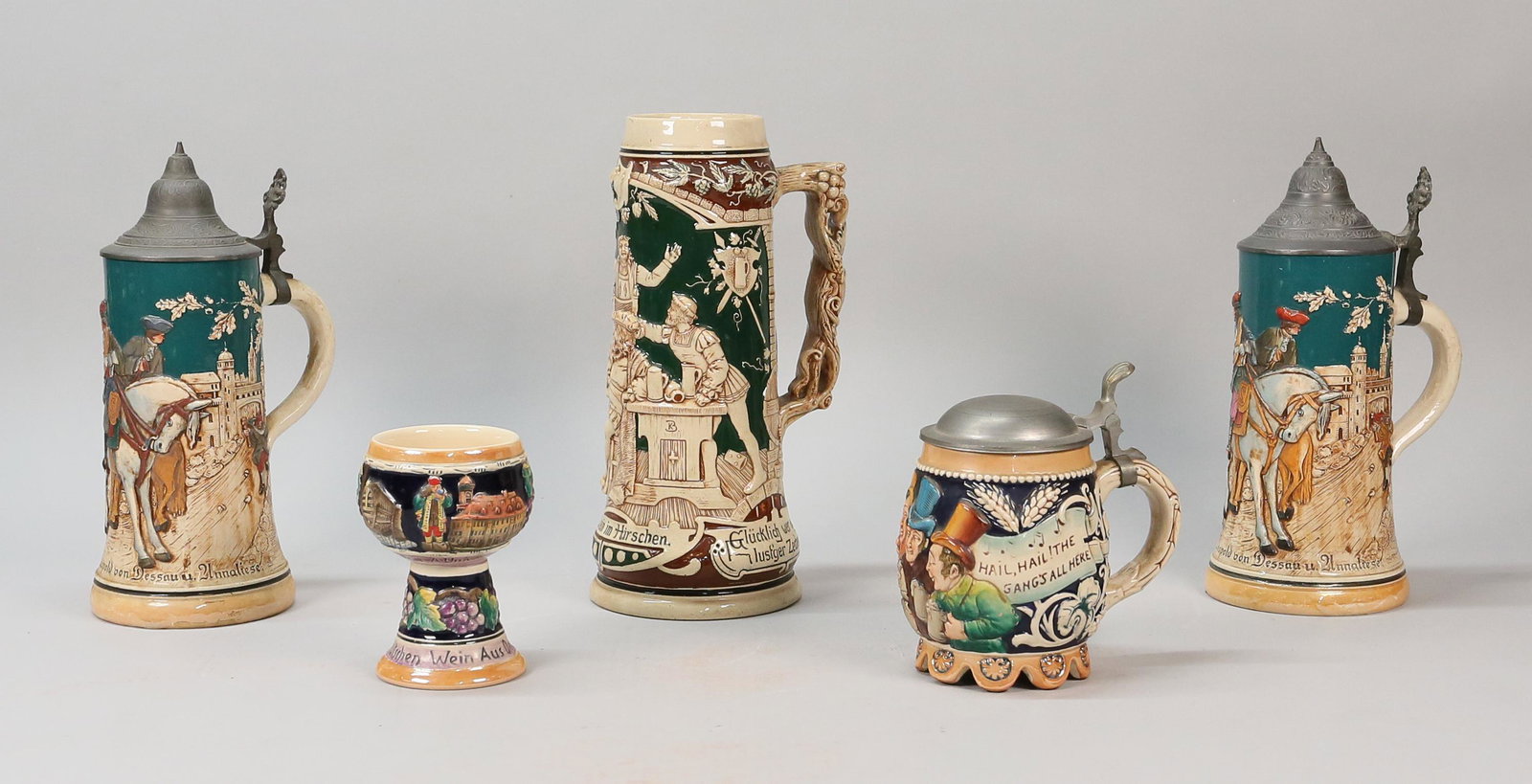 German Porcelain Goblet & 4 Steins: Lot of German porcelain steins and a goblet. Pair of Leopold von Dessau steins, each 11"H including lids; Rodenstein im Hirschen (no lid), 11 1/2"H; goblet, 5 1/4"H; musical stein, 5 3/4"H. Minor roug