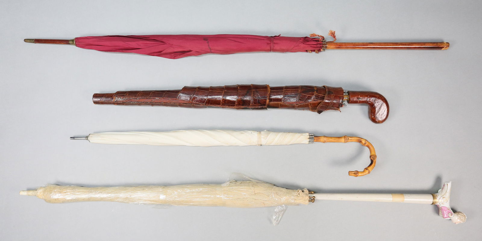 4 Umbrellas & Parasols: Lot of 4 umbrellas and parasols. Painted wood parasol with figural porcelain handle, 38 1/4"; parasol with bamboo form handle, 26 1/2"L; red parasol with wooden handle, 37"L; umbrella with alligator h