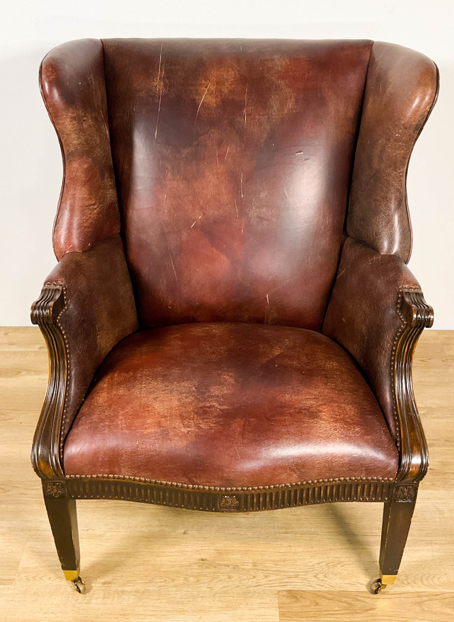Leather Library Wing Chair: Leather library wing chair. With nail heads and carvings. Front feet on brass casters. Leather worn with discoloration and scratches. Wood with rubbing and nicks throughout. 41 1/2" Tall x 33 1/2" Wid