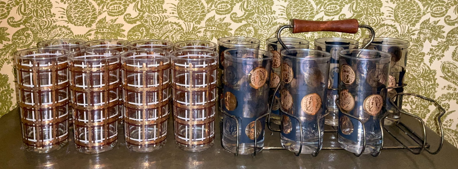 15 Vintage Mid-Century Highball Glasses Carrier: Fifteen vintage mid-century highball glasses. One set of seven glasses (missing one) with coin motifs featuring classical profiles and inscriptions in a wire rack with bronze finish and wood handle. S