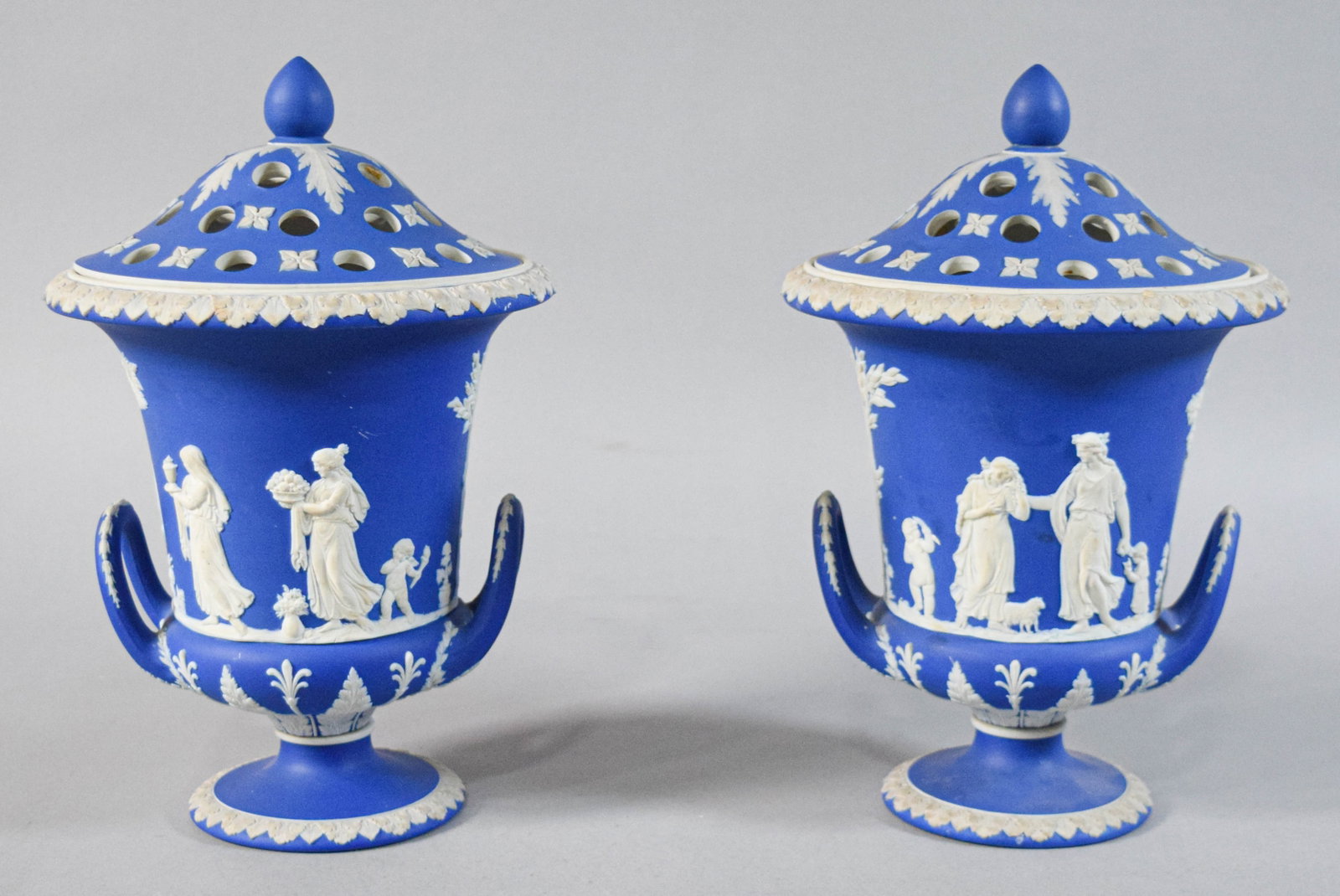 Pair of Wedgwood Cobalt Blue Jasperware Lidded Urns: Pair of Wedgwood cobalt blue Jasperware lidded urns. Domed, perforated lids and twin loop handles. Exterior decorated with continuous neoclassical scenes. Marked Wedgwood, W, KCD on the base. 11" H x 