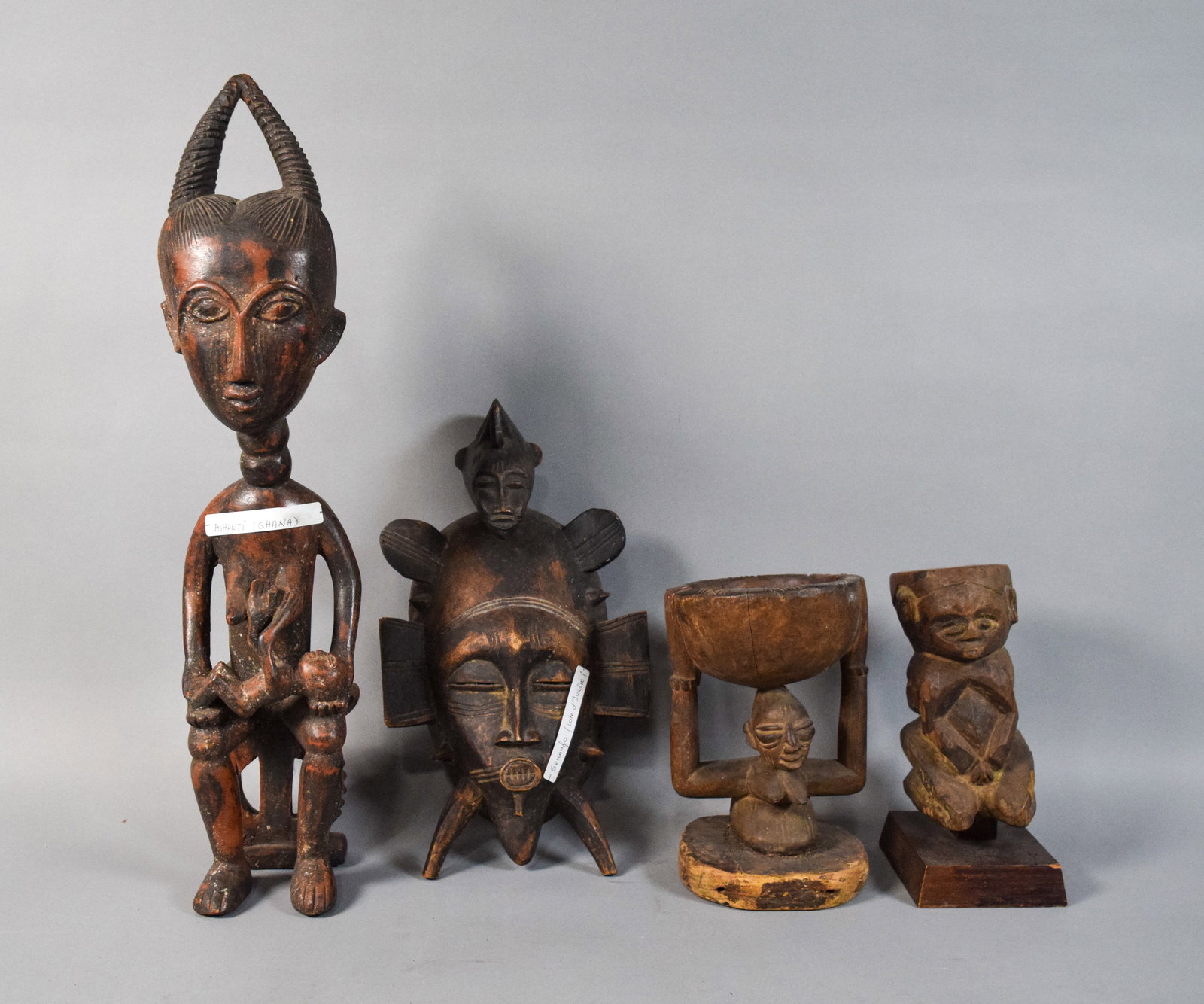 4 West African Style Carved Wood Tribal Figures: Group of four West African style carved wooden objects including a maternity figure, a mask, and two vessel-bearing figures. The maternity figure depicts a seated nursing mother with a paper label rea