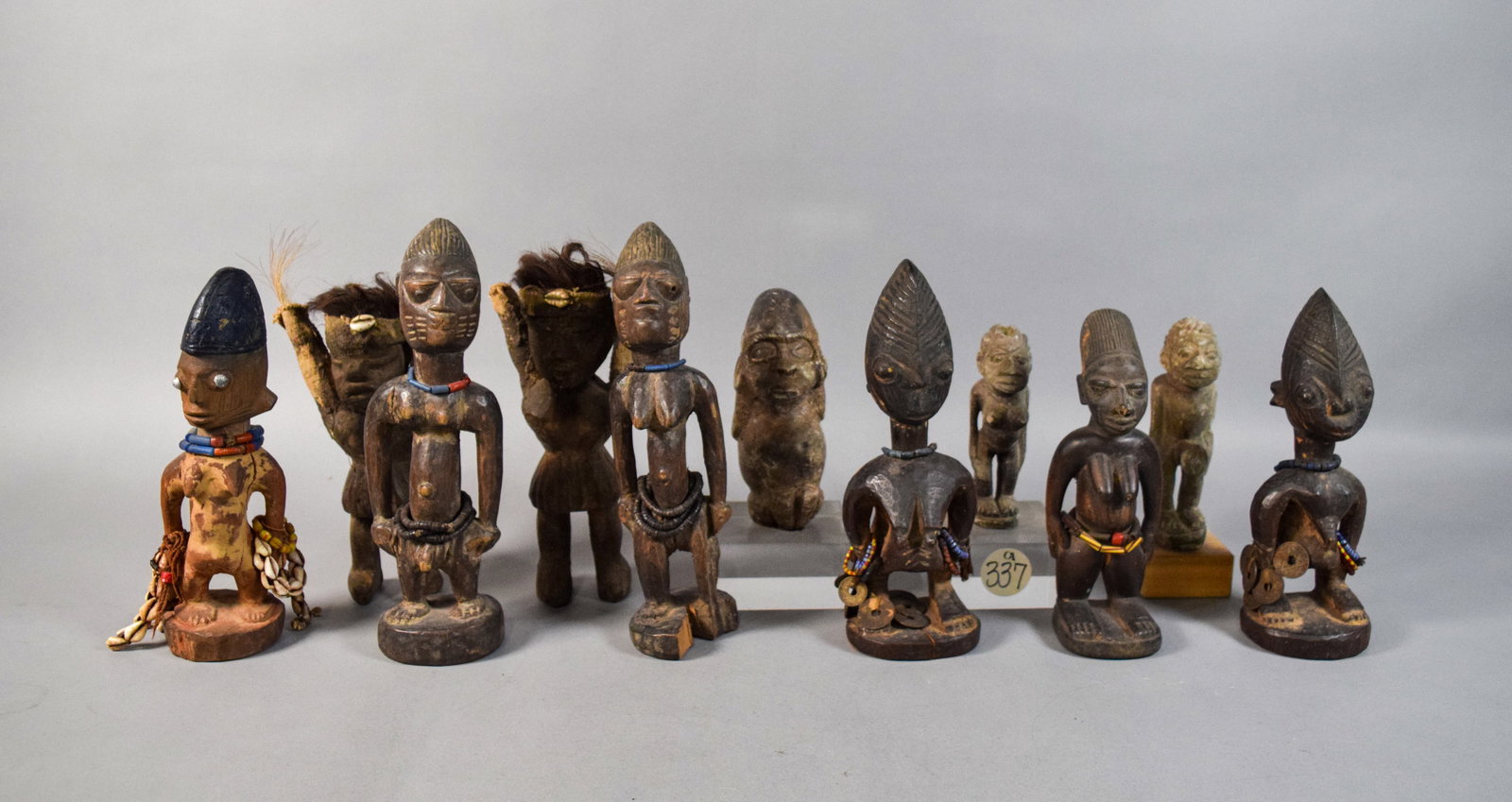11 West African Style Carved Wood Figures: Group of 11 West African style carved wood figures including three smaller figures carved from stone. Several figures adorned with cowrie shells, animal hair, beads, and metal coins. Largest figure 11
