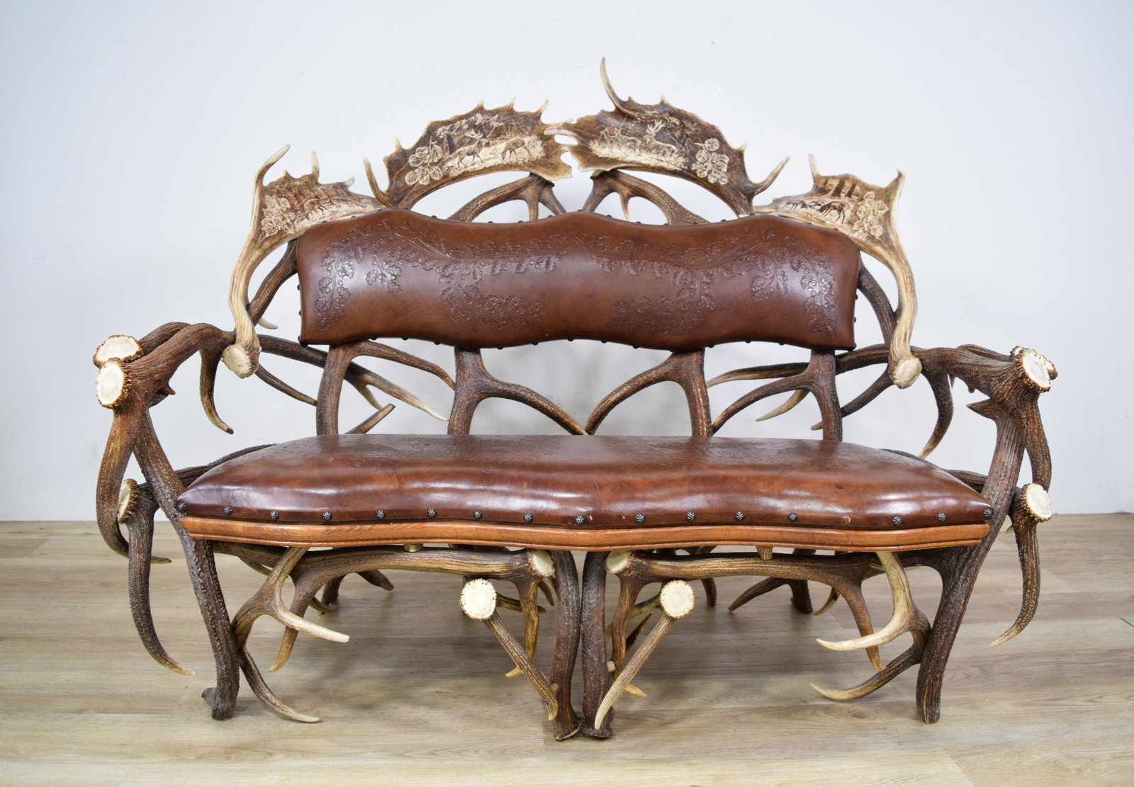 Antler Settee With Tooled Leather Upholstery: Antler settee with tooled leather upholstery. Visible wood frame with interlocked antler base. Upholstered in brown leather, with the backrest featuring tooled oak leaf and acorn scrollwork and the se