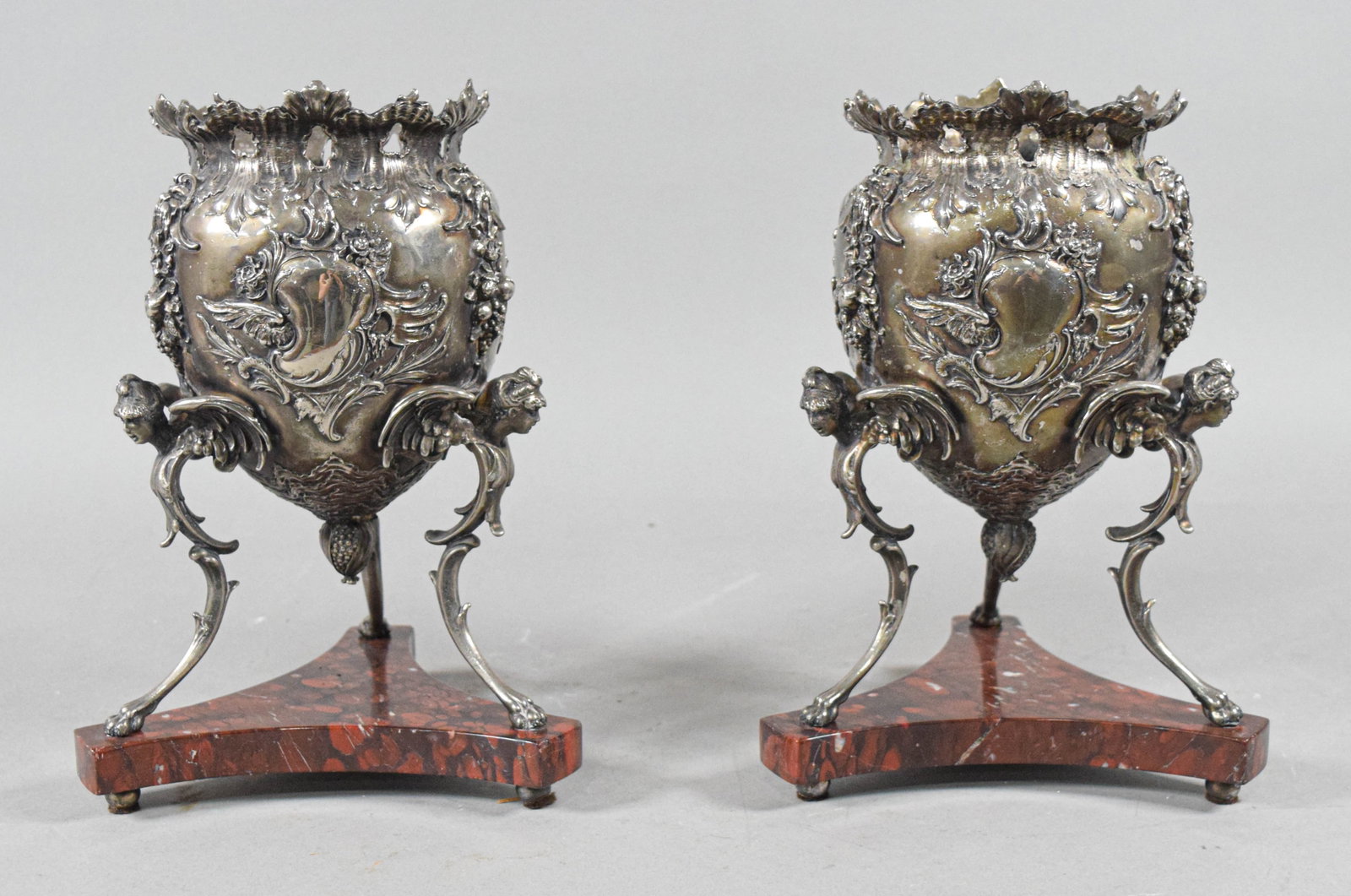 Pair of Victorian Silver-Plated Urns on Marble Bases: A pair of silver-plated urns in the Victorian Neo-Rococo style. Each urn features a bulbous body decorated with high-relief repouss?? floral swags, scrolled cartouches, and acanthus leaves. The flared