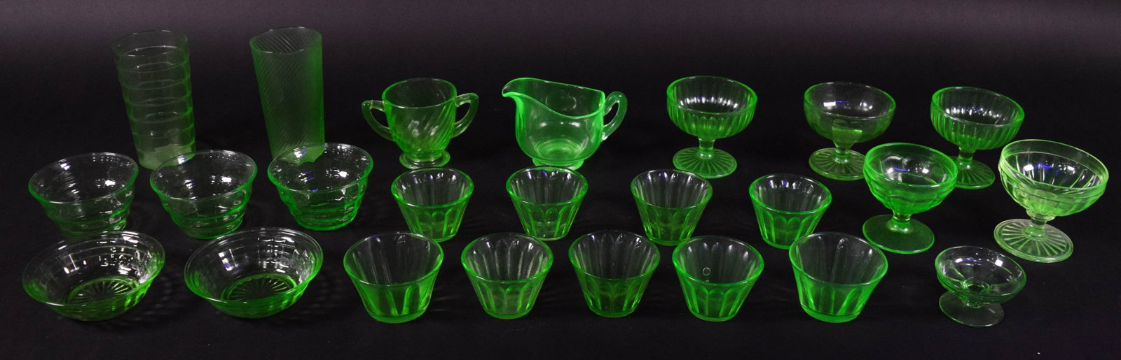 Collection of 20+ Pieces of Uranium Glass Tableware: A collection of 23 pieces of Depression-era uranium glass, also known as vaseline glass, featuring a transparent green color that displays strong neon green fluorescence under ultraviolet light. The a