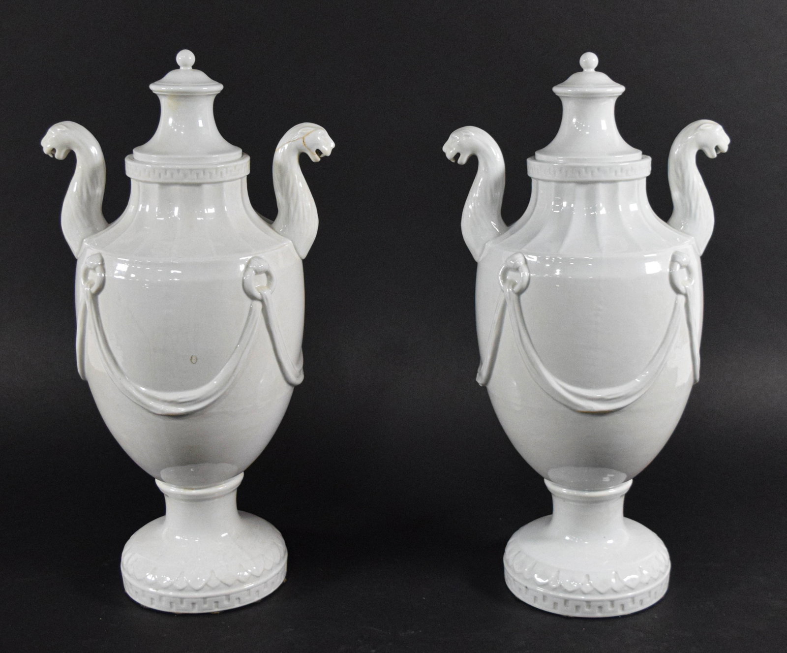 A Pair of Neoclassical Style White Glazed Porcelain Panther Head Lidded Urns: A pair of white glazed porcelain lidded urns in the Neoclassical style. Each urn features an ovoid body decorated with raised relief swags suspended from circular ring motifs. The zoomorphic handles a