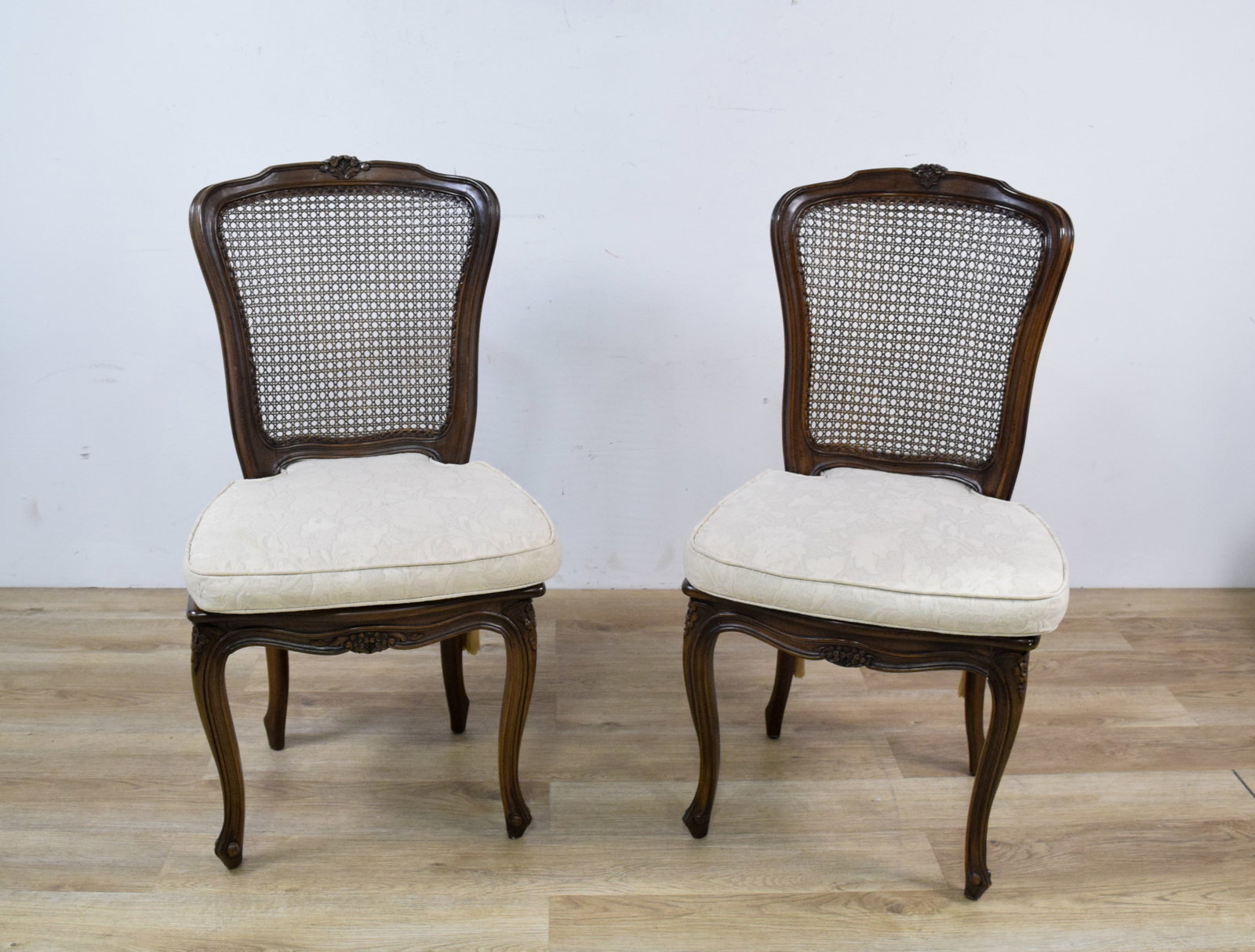 A Pair of Louis XV Style Cane Back Side Chairs: A pair of Louis XV style dining side chairs constructed with a curved wood frame. Each chair features an arched crest rail centered by a carved floral motif. The back panels and seat decks are fitted 