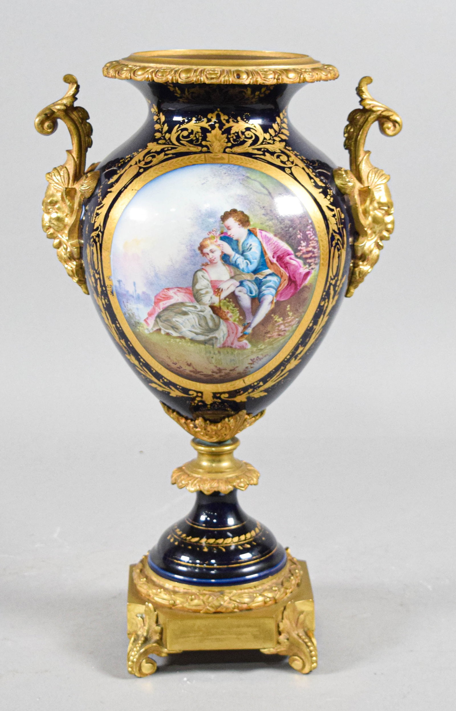 Late 19th Century French Cobalt Blue Porcelain and Ormolu Mounted Urn: Large porcelain decorative pedestal urn featuring a cobalt blue ground. The body is decorated with a hand-painted pastoral scene depicting a couple in 18th-century attire within an elaborate gilt scro