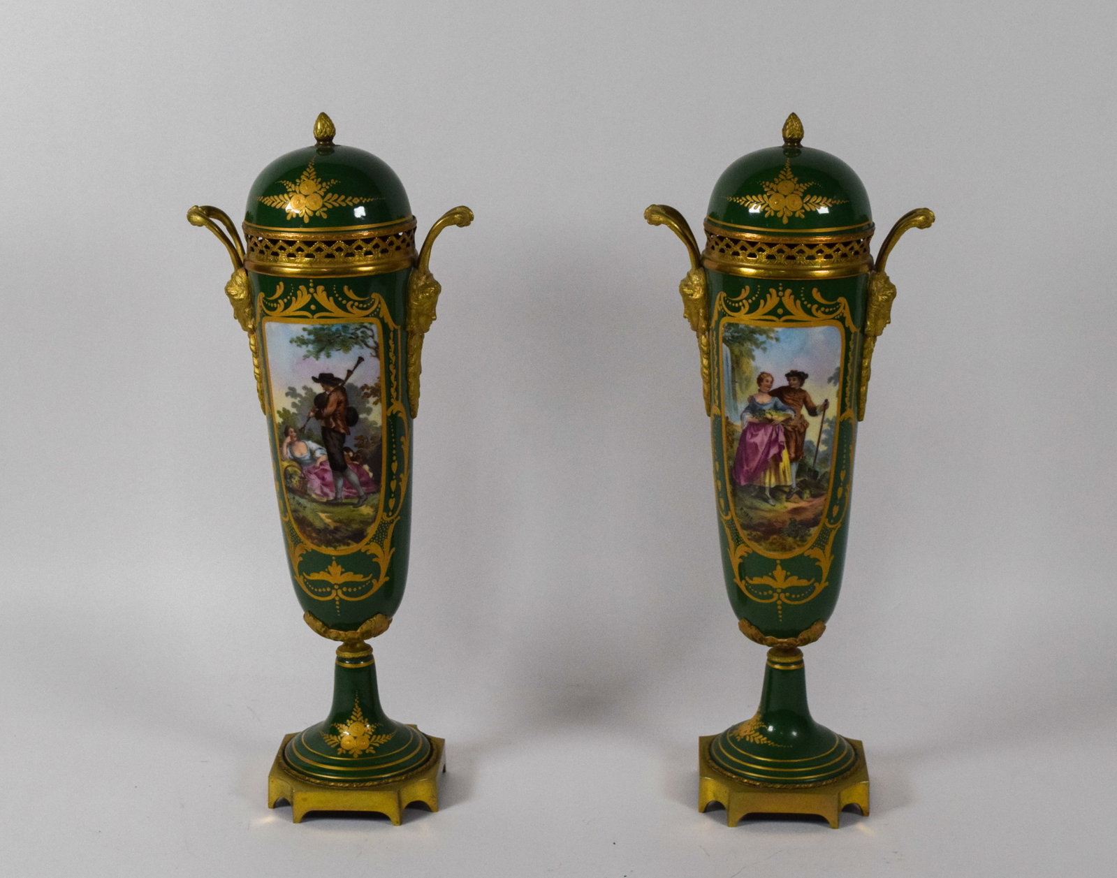 A Pair of Sevres-Style Gilt-Bronze Mounted Porcelain Lidded Urns: A pair of lidded porcelain urns featuring a forest green ground and hand-painted pastoral scenes. Each urn displays a central cartouche within a raised gilt scrollwork border. One scene depicts a coup