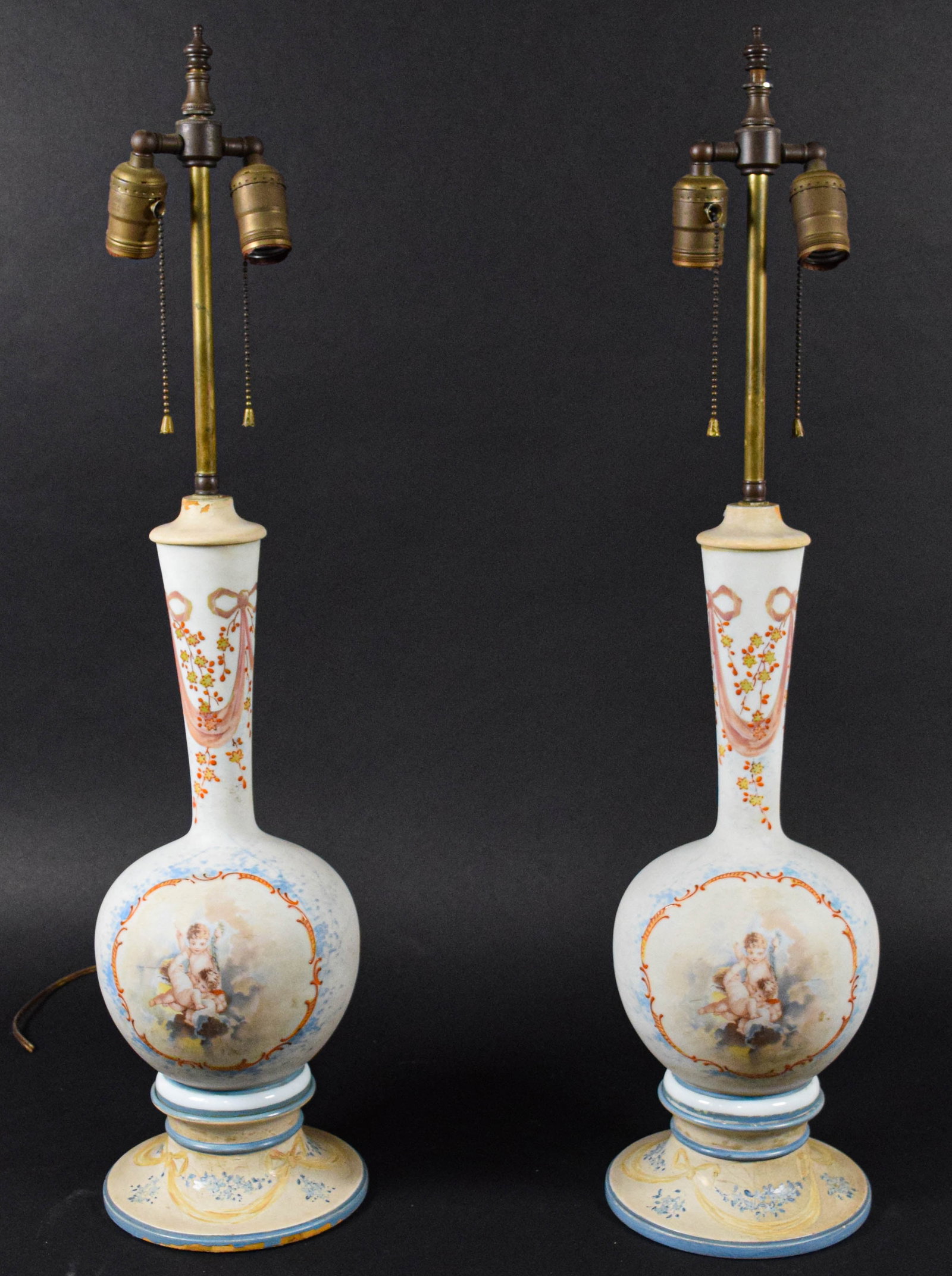 A Pair of Neoclassical Style Porcelain Vase Form Table Lamps with Cherub Decoration: A pair of porcelain table lamps featuring vase form bodies mounted on flared bases. The central bodies are decorated with hand finished scenes depicting three cherubs among clouds, framed by orange sc