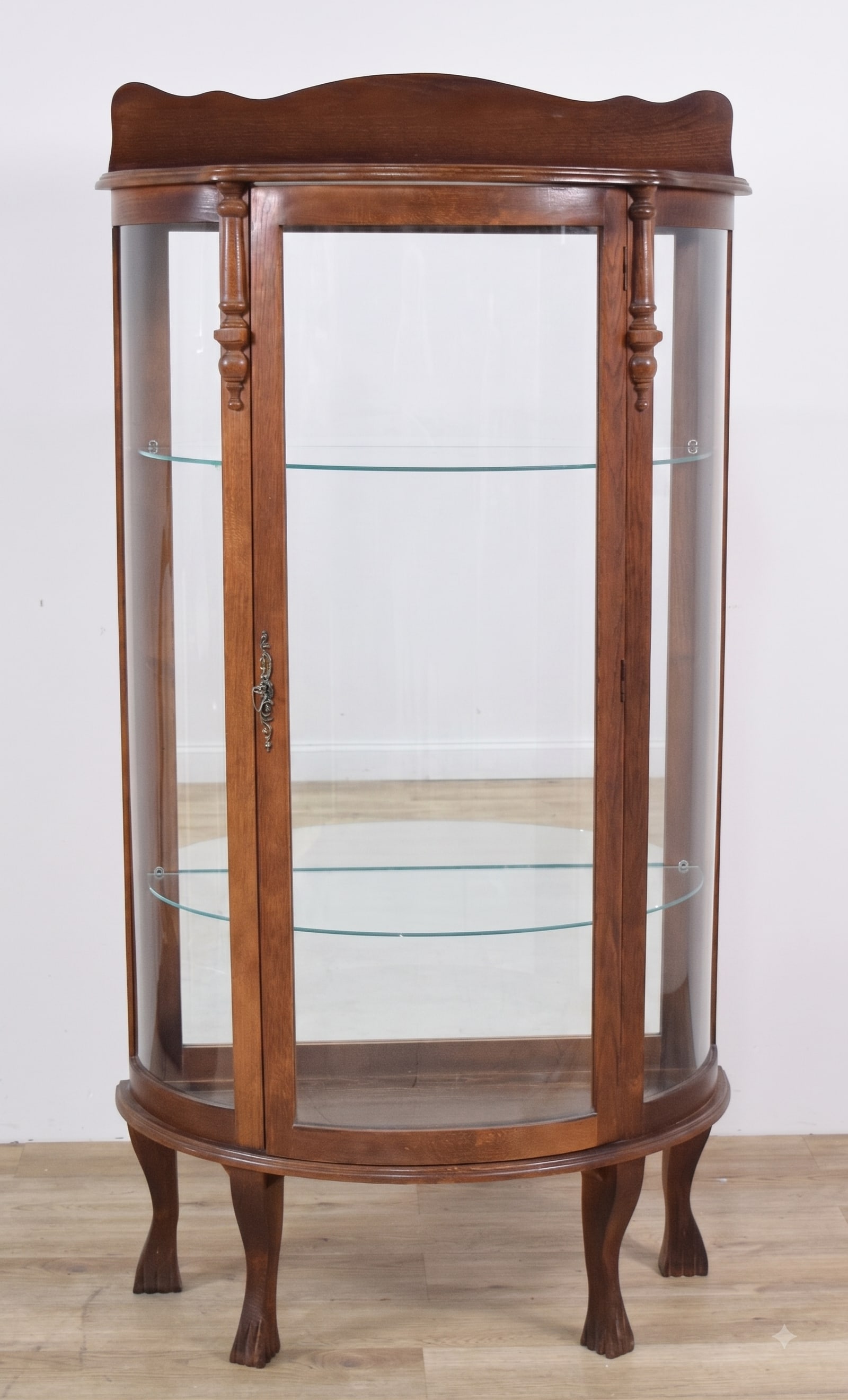 Canadian Oak Curved Glass Display Cabinet with Claw Feet: A curved glass display cabinet of Canadian origin constructed from oak. A single door with an ornate, scrollwork cast-metal escutcheon opens to a mirrored interior back and includes two adjustable gla