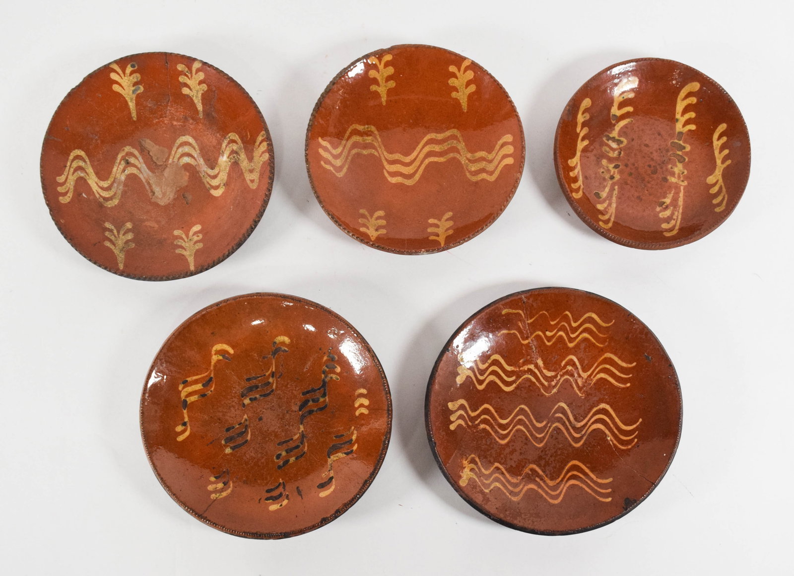 5 American Folk Art Style Slip-Decorated Redware Plates: A group of five slip-decorated redware plates in the American folk art tradition. Each plate is hand-potted with a lead-glazed interior and a characteristic coggled or notched rim. The interior surfac