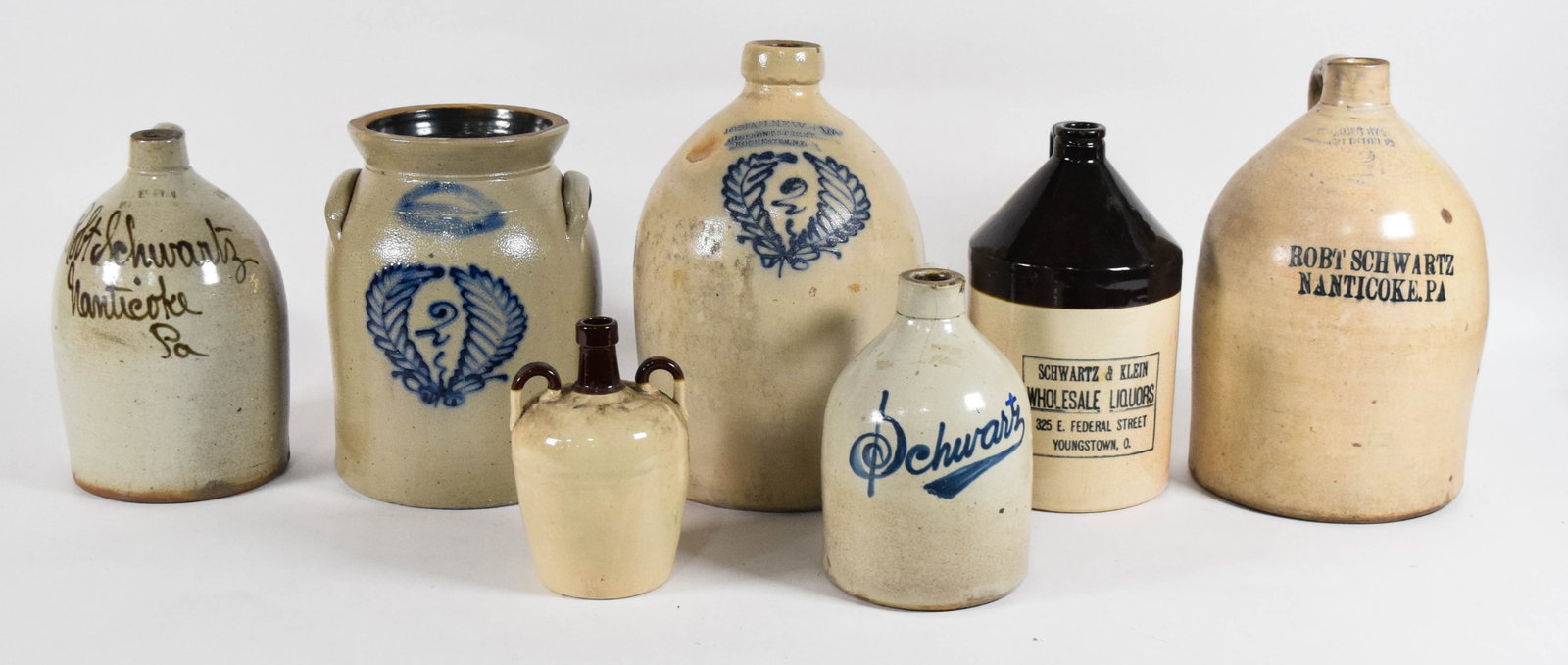 7 American Salt-Glazed and Stenciled Stoneware Advertising Jugs and Crock: An assortment of seven pieces of American stoneware comprising six jugs and one two-handled crock. The collection features late 19th to early 20th-century salt-glazed vessels with regional merchant ad