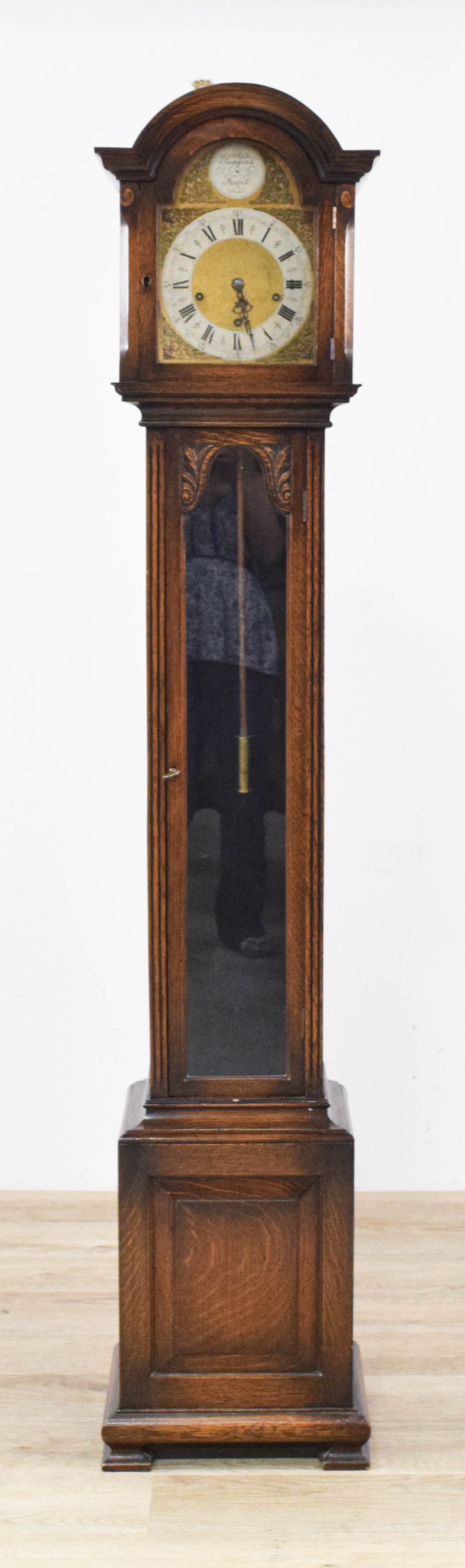 English Oak Grandmother Clock with Tempus Fugit Brass Dial: An English grandmother clock featuring a mechanical two-train movement and a dark-stained oak case. The architecture consists of an arched hood above a slender trunk with a glazed door featuring carve