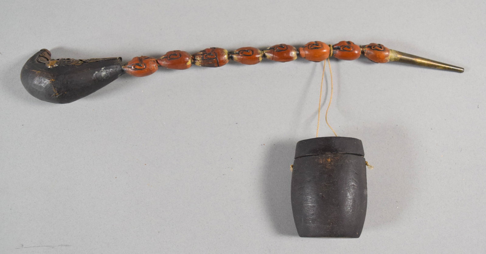 Japanese Carved Wood Opium Pipe with Matching Container: This Japanese opium pipe features a long stem composed of nine carved, reddish-orange beads. Each bead is hand-carved with stylized motifs that appear to be calligraphic or floral in nature. The pipe'