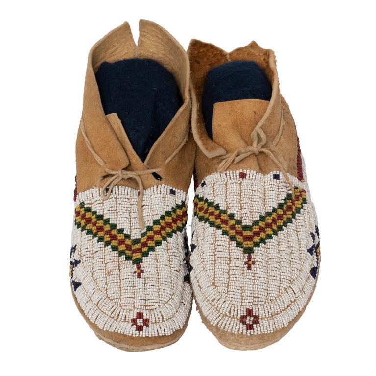 Pair of Beaded Moccasins: Two-piece tanned leather construction with hard soles. Beaded with green, red, yellow, and blue geometric designs on a white background. Square stitched vamps. **Very good condition overall. Some disc