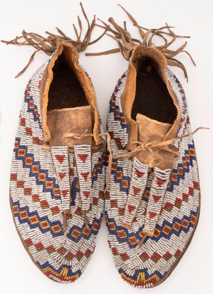Pair of Plains Beaded Moccasins: Two-piece tanned hide construction with sinew-sewn hard soles and split sewn-on tongues. Rear seam with heel fringe. Beaded throughout with red, blue, gold, white, and Yellow beads in a geometric moti