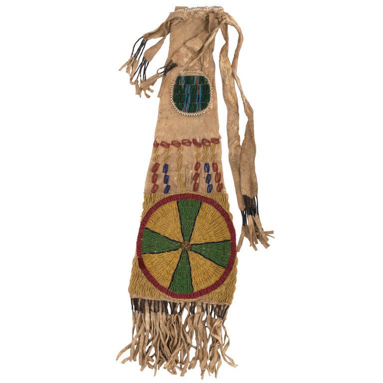 Beaded Pipe Bag: 18.5" x 6.6" without tassels. Buckskin construction with quill-wrapped tassels and attached pouch. Beaded throughout with red, green, yellow, and blue beads with a large wheel motif. Attached pouch be