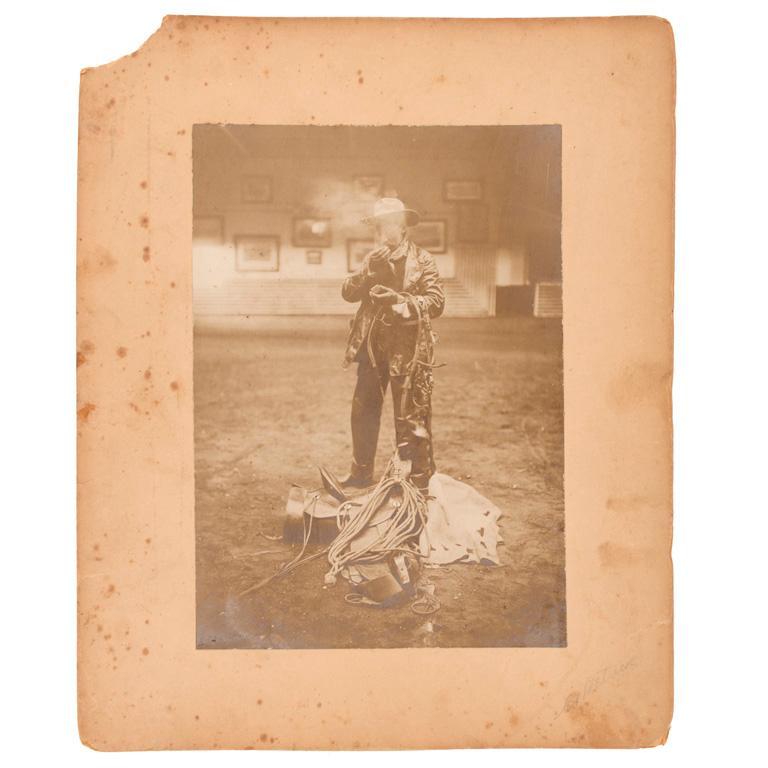 Large Format Photograph of a Cowboy: Mount measures 9" x 11". Large format photograph depicting a cowboy with saddle and horse tack, lighting a cigarette. Appears to be an interior shot, probably at an indoor rodeo or barn. **Some wear t