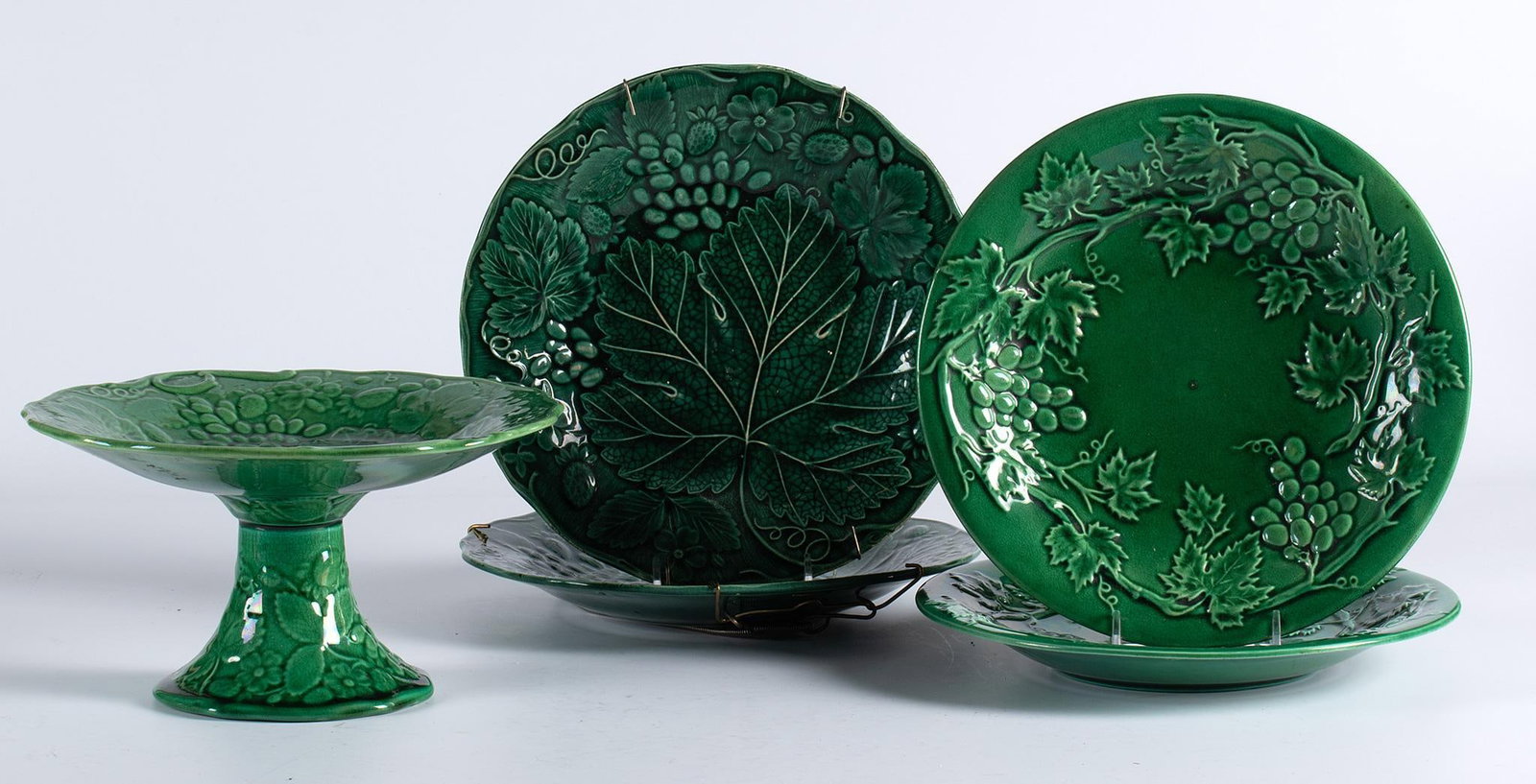 A Group of Foliate Decorated Majelica: American, late 19th century. A grouping of four plates and a single compote majelica tablewares, each with central leaf and overall foliate fruit design; largest ht. 5, dia. 9 inches. Overall good con