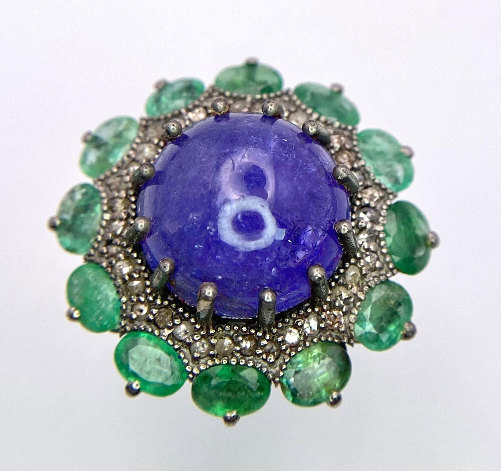 A 11.80ct Tanzanite Cabochon Gemstone Ring with halos of 3.5ctw Emeralds and 0.70ctw Diamonds. Set: A 11.80ct Tanzanite Cabochon Gemstone Ring with halos of 3.5ctw Emeralds and 0.70ctw Diamonds. Set in 925 silver. Size M. 10.52g total weight. Comes in a presentation case and GLI certificate.Ref: CD-