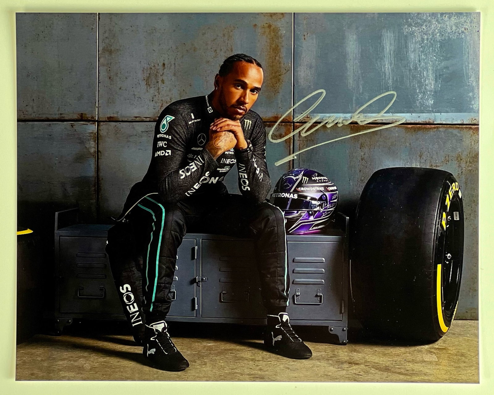 Lewis Hamilton- F1 Champion SIgned Photograph: Autographed 8" x 10" photograph of Formula 1 champion Lewis Hamilton. The image showcases Hamilton in his Mercedes racing suit, seated confidently beside a racing tire and helmet, exuding the calm int