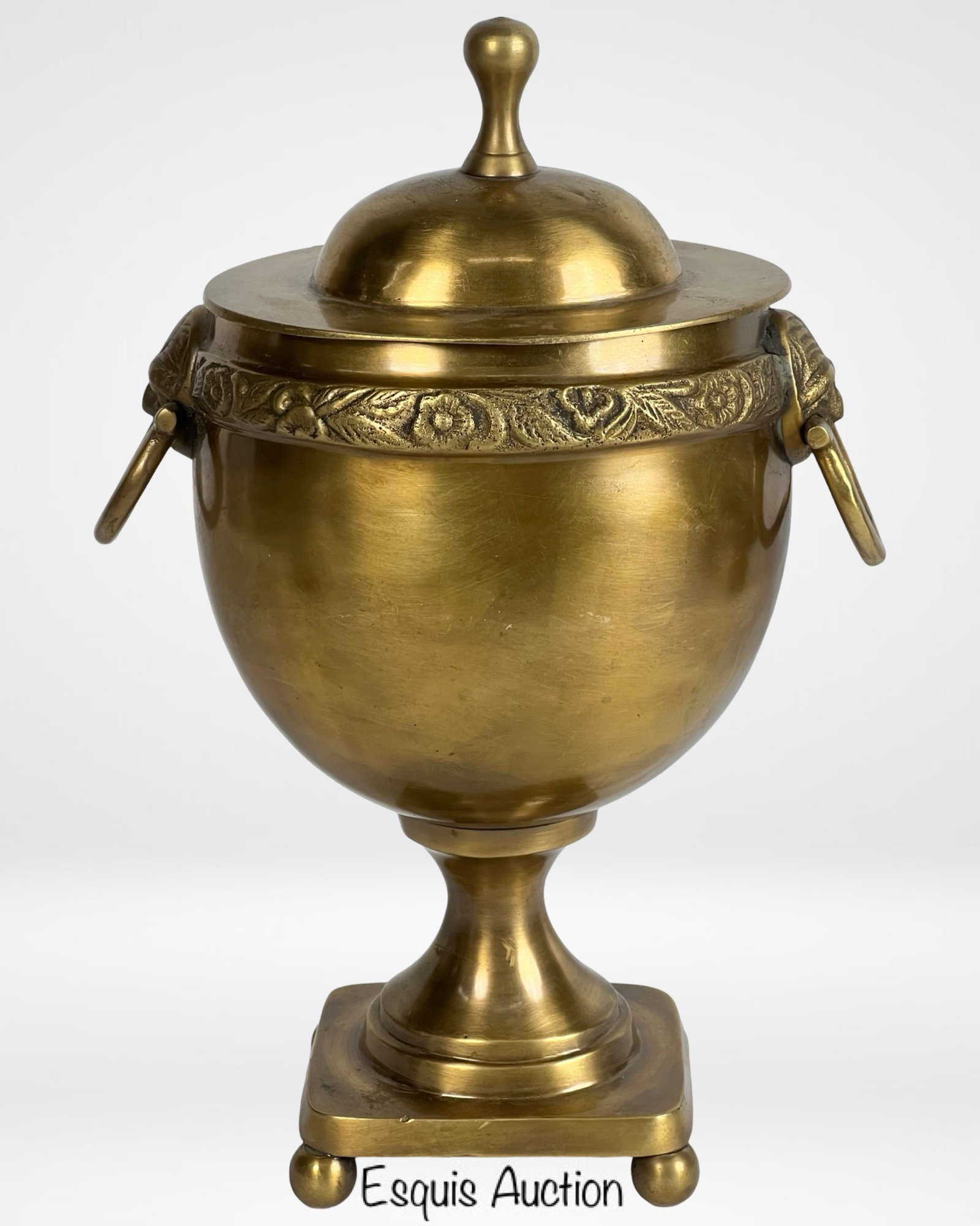 Mottahedeh Brass Urn with Lion Head Handles: A classic Mottahedeh brass urn featuring a lidded design with decorative lion-head handles and floral detailing around the rim. This elegant piece rests on a square pedestal base with ball feet, combi