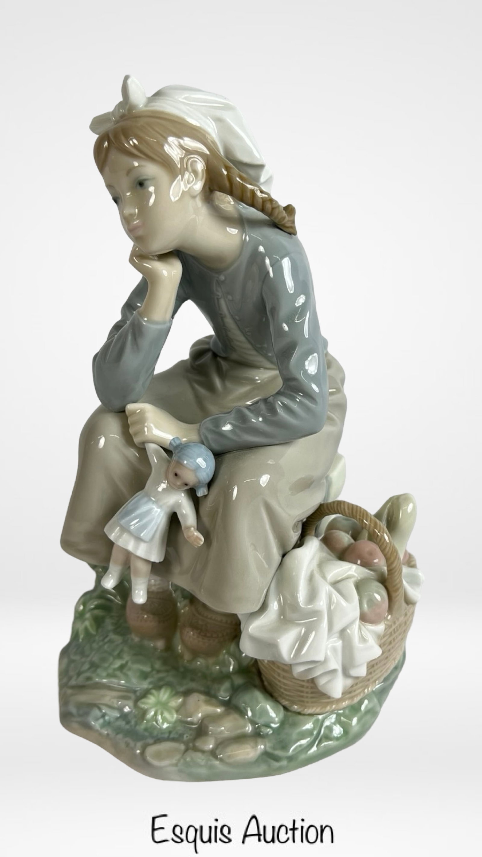 Lladro "Girl with Doll" Porcelain Figurine: A charming Lladro porcelain figurine titled "Girl with Doll," crafted in 2011. This piece depicts a young girl seated pensively on a rock with a delicate porcelain doll in hand, surrounded by a basket