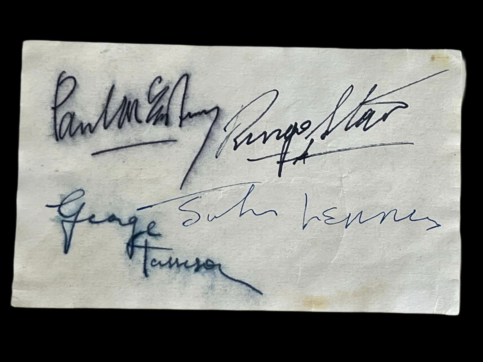The Beatles - Fully Signed Autograph Card: Autograph card signed by all four members of The Beatles-John Lennon, Paul McCartney, George Harrison, and Ringo Starr. The signatures are on a 3" x 5" tan-colored paper, with Lennon signing in blue b