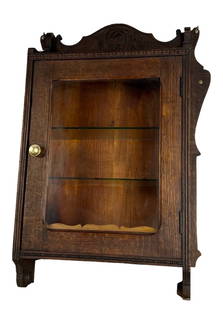 Antique Carved Oak Wall Cabinet with Glass Door: Elegant antique oak wall cabinet featuring a glass door framed in finely grained wood and topped with a carved crest bearing decorative scrollwork. The interior includes two glass shelves and a