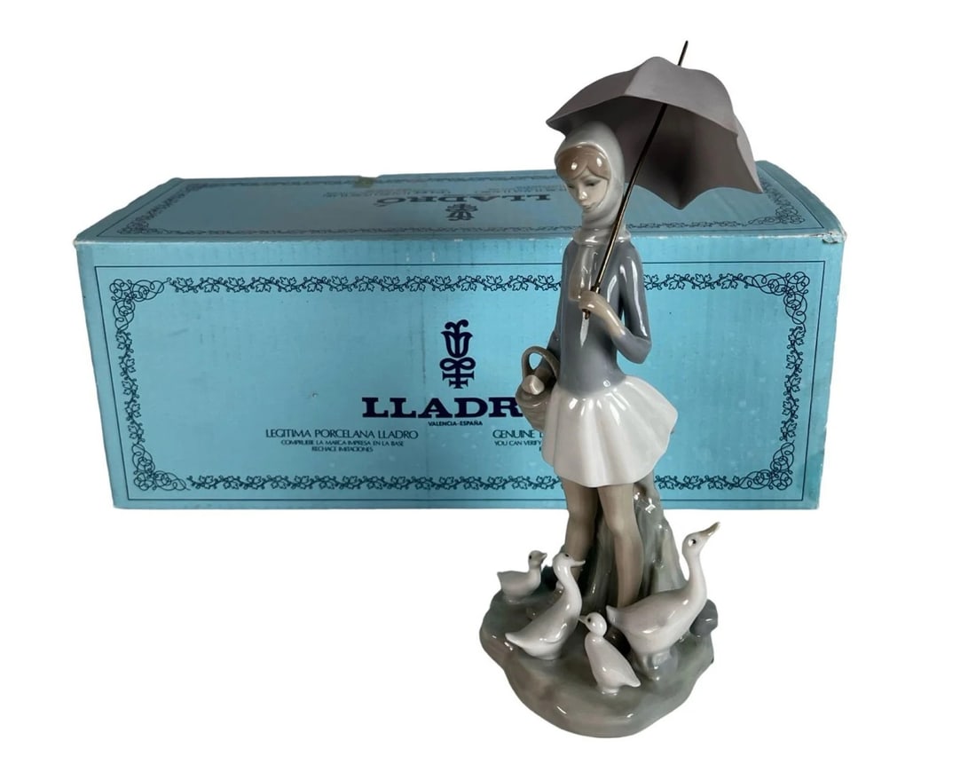 Lladro Girl w/ Umbrella & Geese Porcelain Figurine: Charming and serene, this glossy porcelain figurine by Lladro depicts a young woman holding an umbrella, standing amidst a gentle gathering of geese. Titled "Girl with Umbrella and Geese" and model