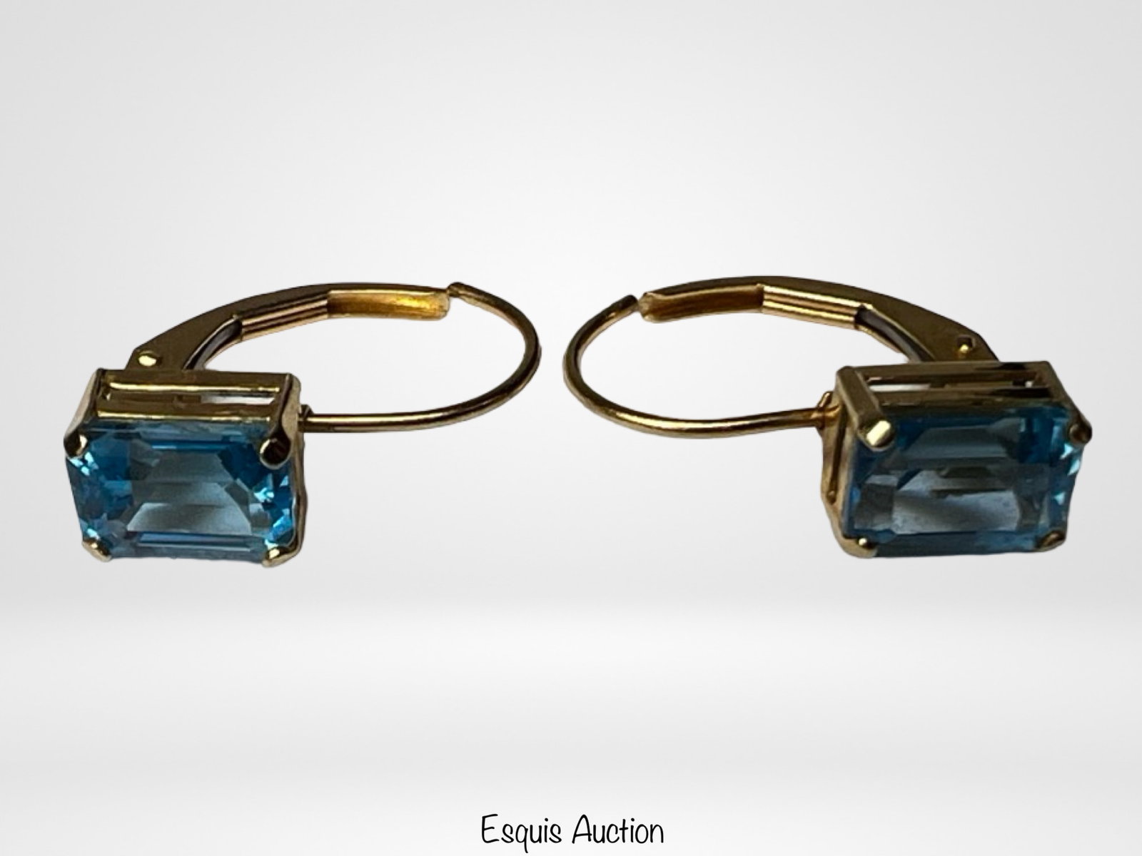 14k Gold Leverback Dangle Blue Topaz Earrings: Elegant pair of 14k yellow gold leverback dangle earrings featuring emerald-cut blue topaz gemstones in open-back four-prong settings. The vivid sky-blue stones are beautifully faceted, offering excep