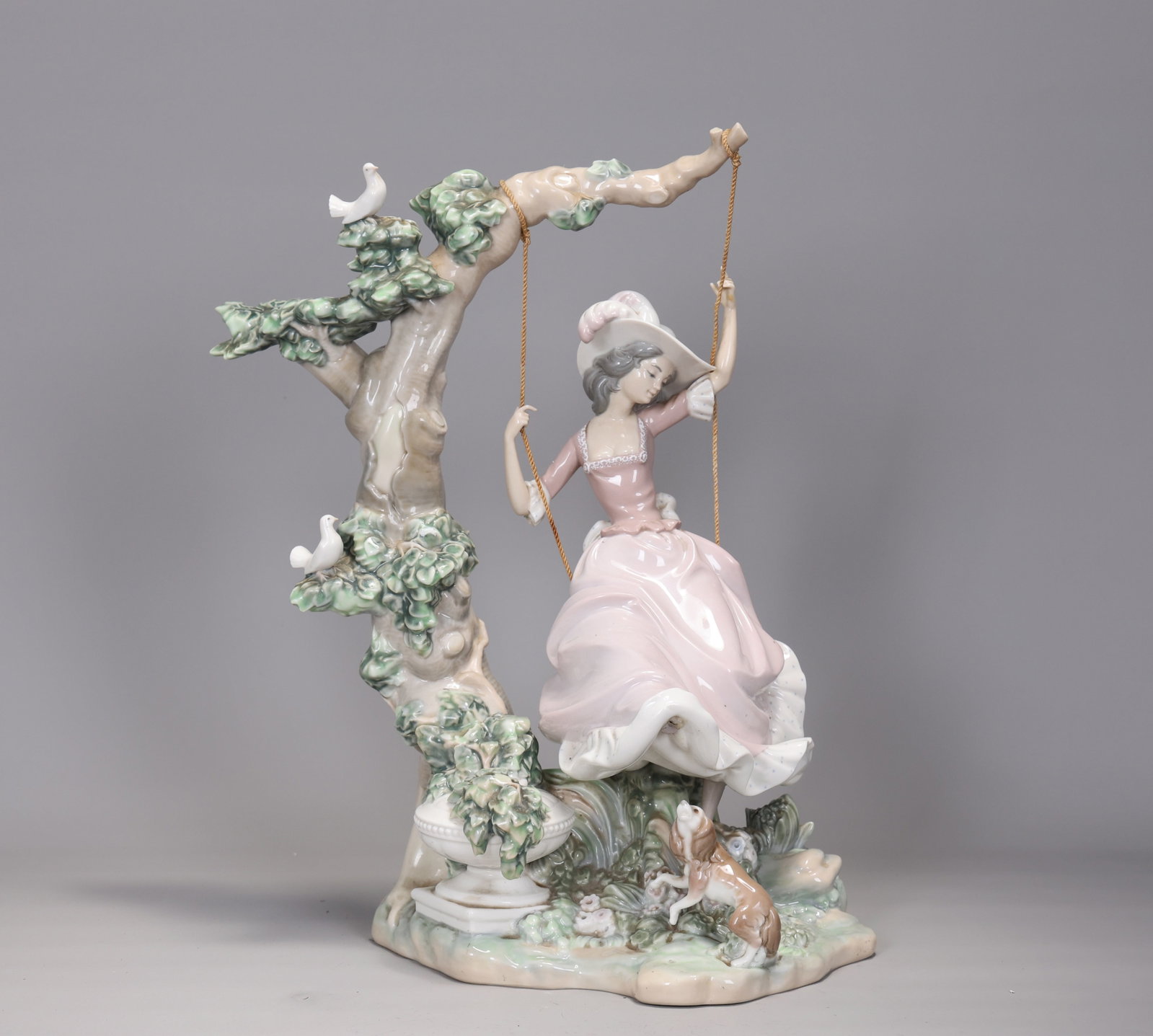 Lladro #1297 Victorian Girl On Swing: Lladro #1297 Victorian Girl On Swing(43 x 30 x 20 cm)Composed of glossy glazed porcelain depicting a woman on a swing with a large tree, bird, and dog motifs surrounding her. Lladro backstamp.This pie