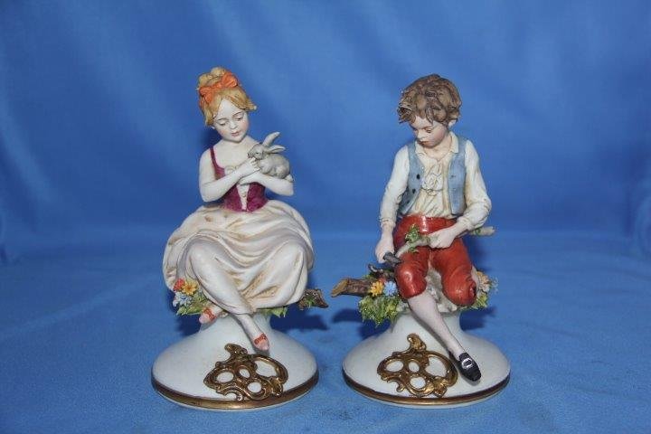 Pair of Capodimonte Figures: 24-4284 Pair of Capodimonte figures. Boy and a girl sitting on a log with flowers. One small chip on the girl's foot.