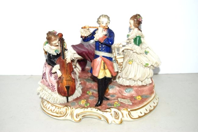Dresden Musical Trio Figurine: 151-5198 Dresden figurine depicting woman playing piano with sheet music, man standing and playing the flute, and seated woman playing the cello; figurine measures 7" tall, 9" wide, and 7" long, marke