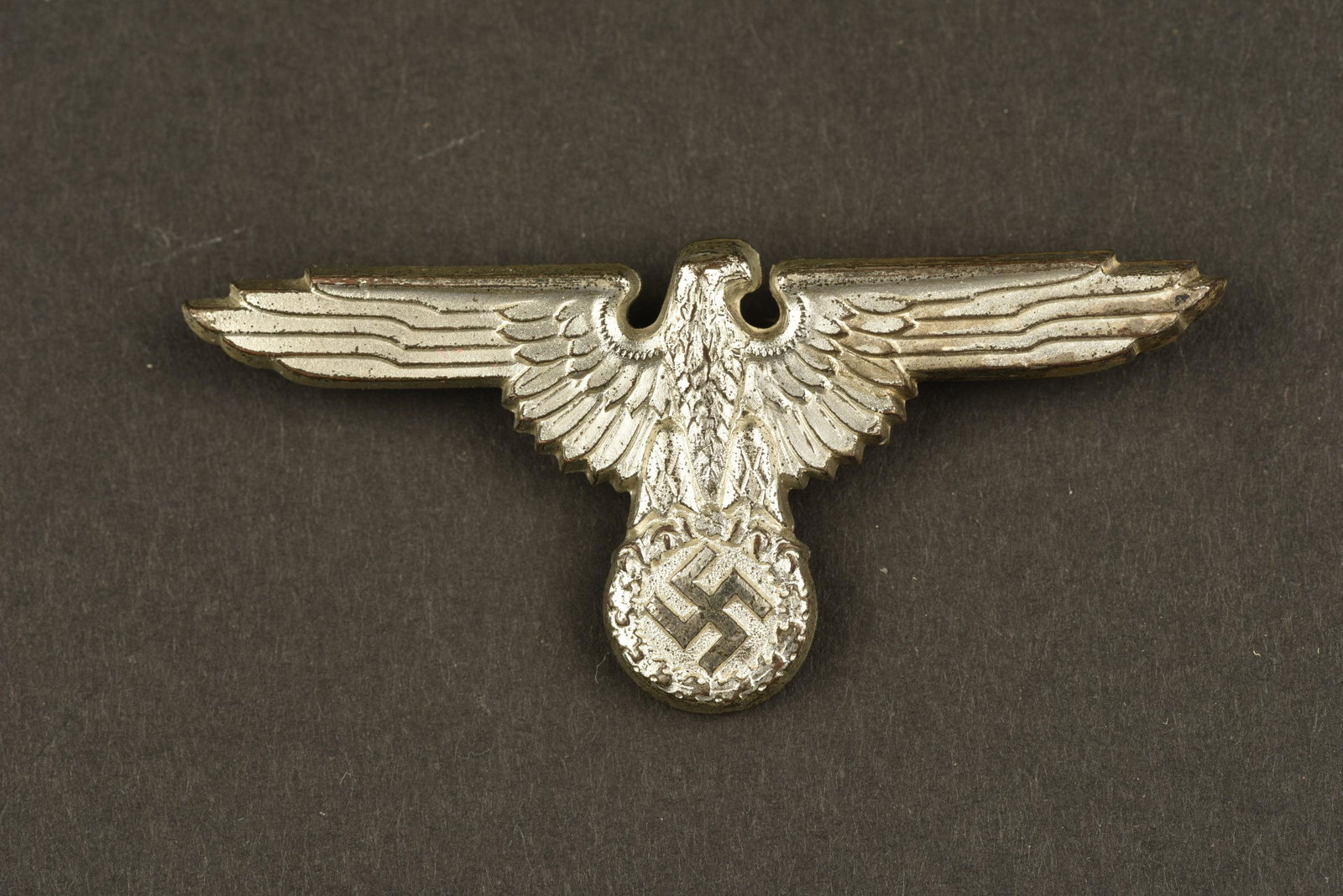 Aigle de casquette de la Waffen SS. Waffen SS cap eagle: En aluminium, les pattes de fixation sont presentes. Couleur argente Ã 80%. Marquages presents, mais illisibles. Certainement une fabrication Ferdinand Wagner. A noter une certaine usure et patine 