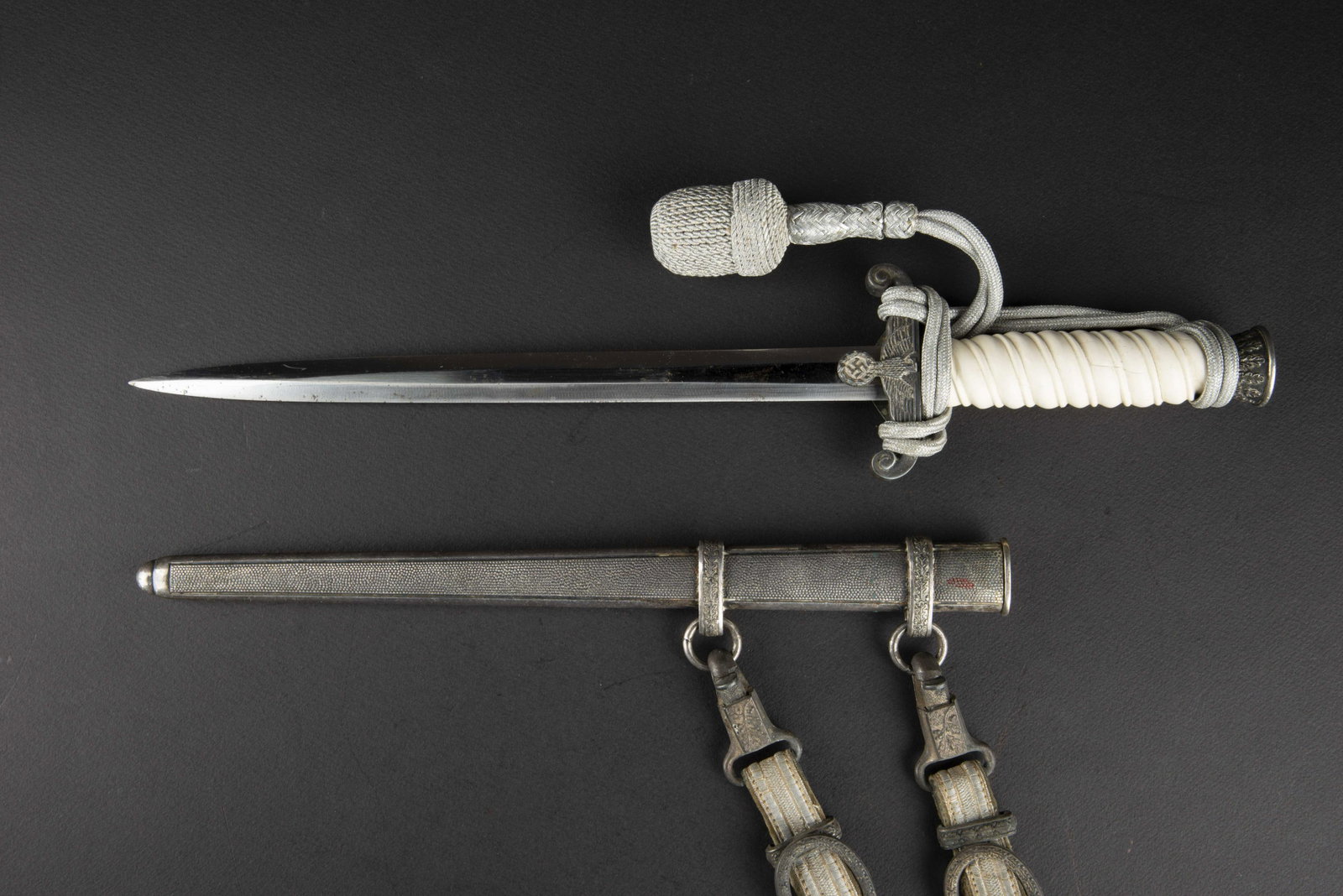 Dague d officier de la Heer. Heer officer dagger.: Poignee en celluloi d blanche, nombreuses fissures. Dragonne en fil d argent montee de maniere reglementaire. Garnitures en metal mat. Lame complete dans sa longueur, presentant quelques traces d oxyd
